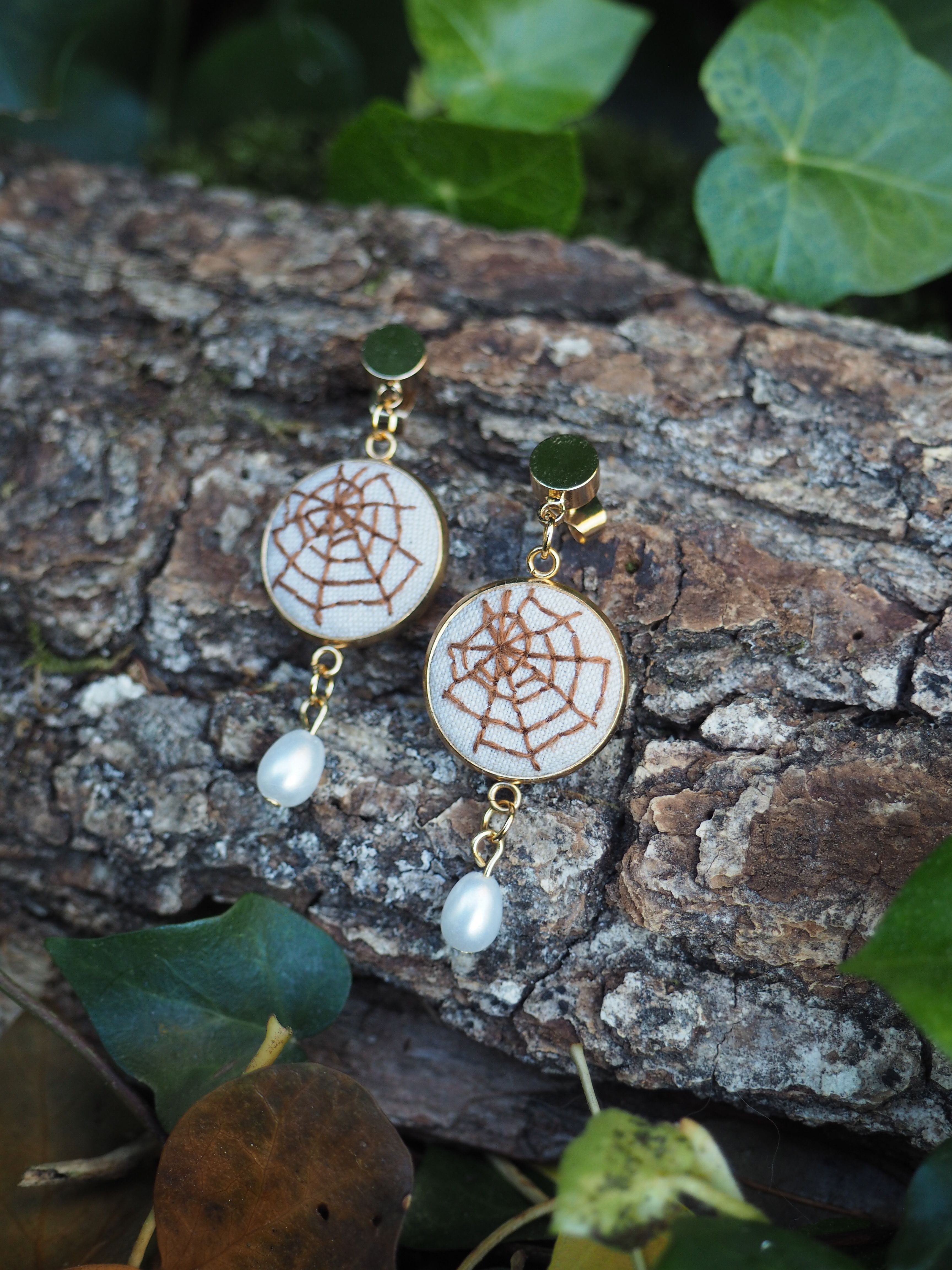 Cette image présente une paire de boucles d'oreilles élégantes disposées sur un morceau d'écorce naturelle dans un environnement végétal. **Description des bijoux :**
- Boucles d'oreilles pendantes avec des crochets dorés
- Pendentifs principaux circulaires de couleur blanche/crème avec un motif décoratif brun rappelant une toile d'araignée ou un mandala
- Petites perles blanches nacrées suspendues en bas de chaque boucle d'oreille **Mise en scène :**
- Les bijoux reposent sur un morceau d'écorce rugueuse aux tons bruns et gris
- L'arrière-plan est composé de feuillage vert, créant un contraste naturel
- L'éclairage met en valeur les détails des boucles d'oreilles et crée une ambiance douce Cette présentation met en valeur le style artisanal et naturel des bijoux, soulignant leur élégance dans un cadre organique qui complète parfaitement leur esthétique.