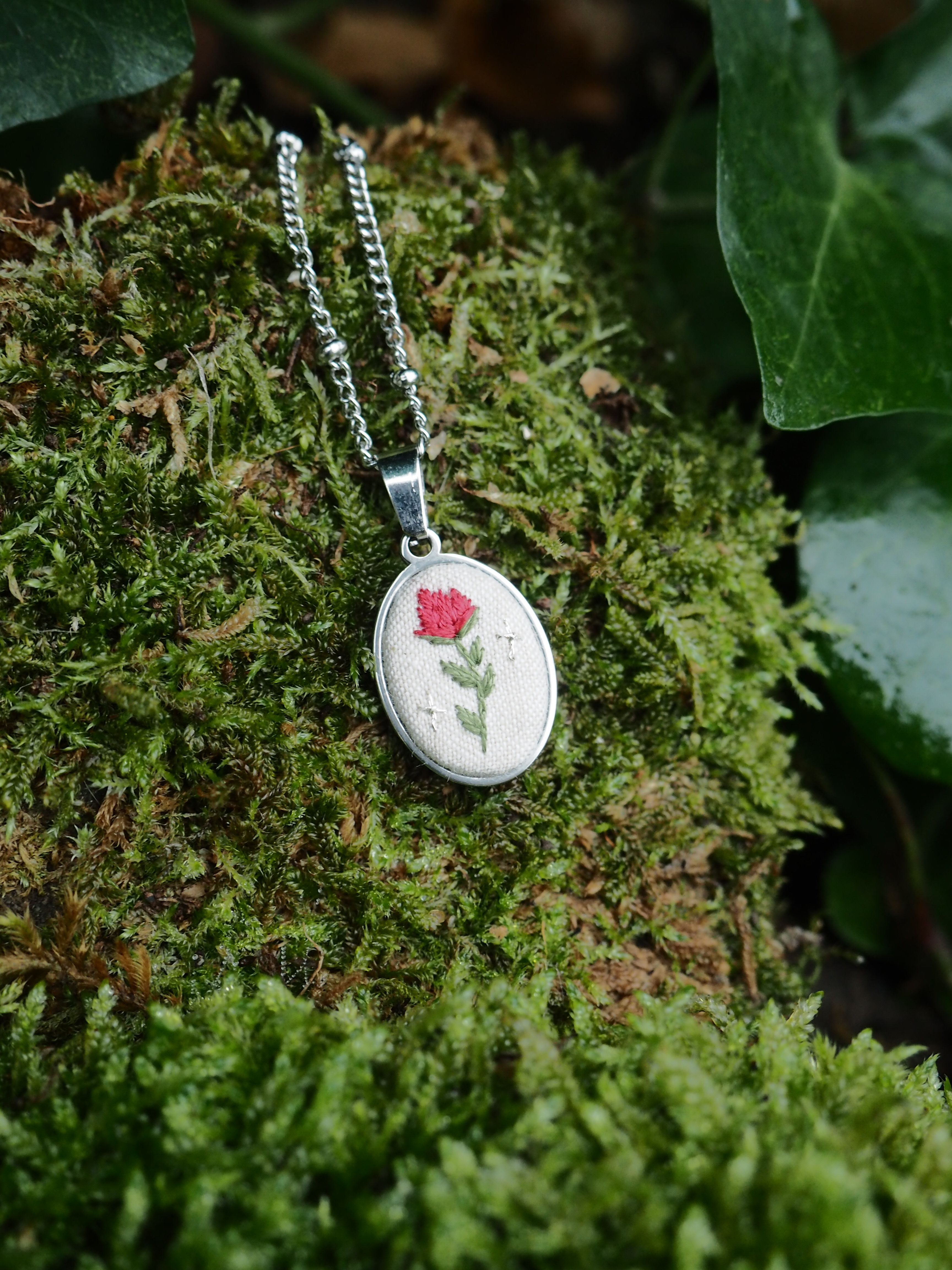 This image features a close-up shot of a delicate, oval-shaped pendant necklace resting on a bed of vibrant green moss. The pendant itself has a silver-toned metal frame, and within it, there's a light-colored, possibly cream or off-white, fabric background. Embroidered on this fabric is a charming design: a small, red flower with a green stem and leaves. To the right of the flower, there are two tiny, white embroidered star-like shapes, adding a whimsical touch. The pendant is attached to a thin, silver-toned chain, which is partially visible, extending upwards and to the left. The moss provides a natural and textured backdrop, with varying shades of green and some hints of brown underneath, suggesting depth and an organic environment. In the upper right corner and along the top edge, darker green leaves, possibly ivy, are visible, further enhancing the natural setting. The lighting appears soft, highlighting the details of the embroidery and the texture of the moss.