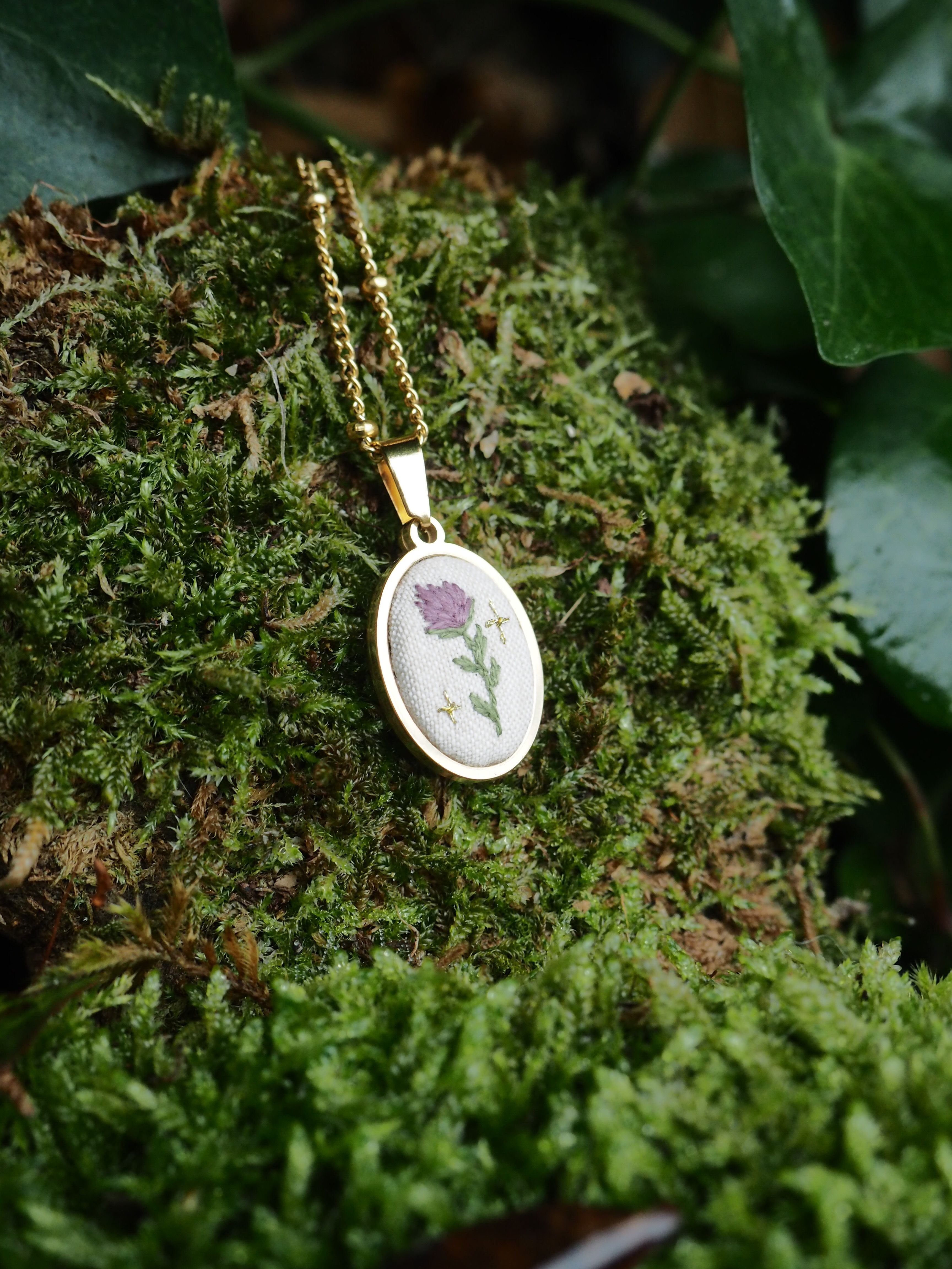 This close-up shot features a delicate gold-colored necklace with an oval pendant, resting on a bed of vibrant green moss. The pendant has a white fabric insert with a small, intricately embroidered design of a purple flower with a green stem and leaves, flanked by two tiny golden star-like accents. The gold chain of the necklace is visible, leading up from the pendant. The moss provides a soft, natural backdrop, with varying textures and shades of green, suggesting an outdoor or natural setting. In the background, hints of darker green foliage, possibly ivy, can be seen, adding depth to the image. The lighting appears soft, highlighting the details of the necklace and the texture of the moss.