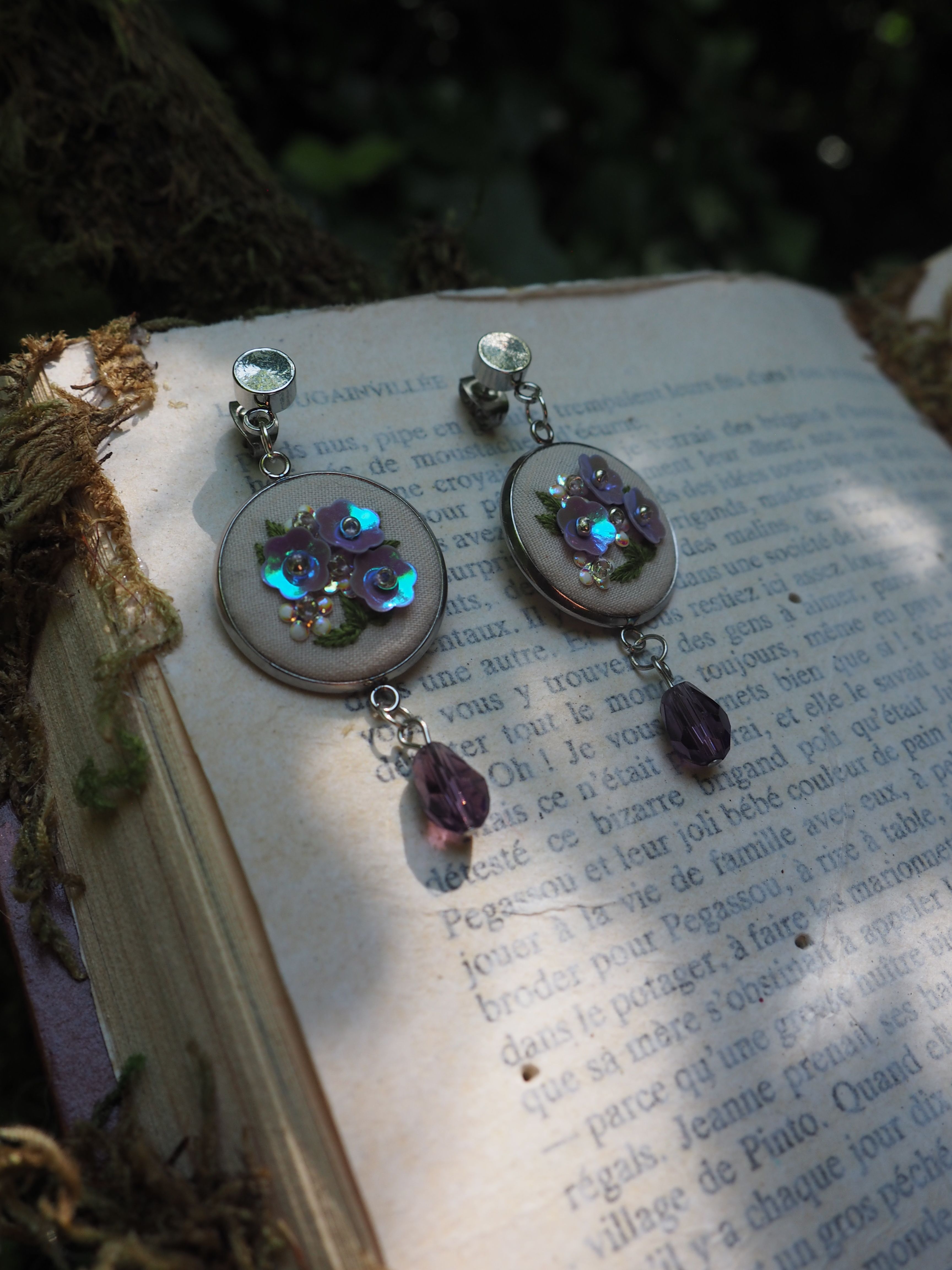 L'image montre des bijoux ou des pendentifs décoratifs posés sur un livre ouvert. Ces pendentifs semblent être faits avec des petites LED bleues qui brillent, créant un effet lumineux mystérieux. Ils sont accompagnés de ce qui semble être des perles ou des cristaux violets. Les pendentifs reposent sur les pages d'un livre en français, dont on peut apercevoir le texte imprimé en dessous. L'ensemble crée une atmosphère féerique et magique, comme si ces bijoux lumineux étaient des artefacts enchantés sortis d'un conte.