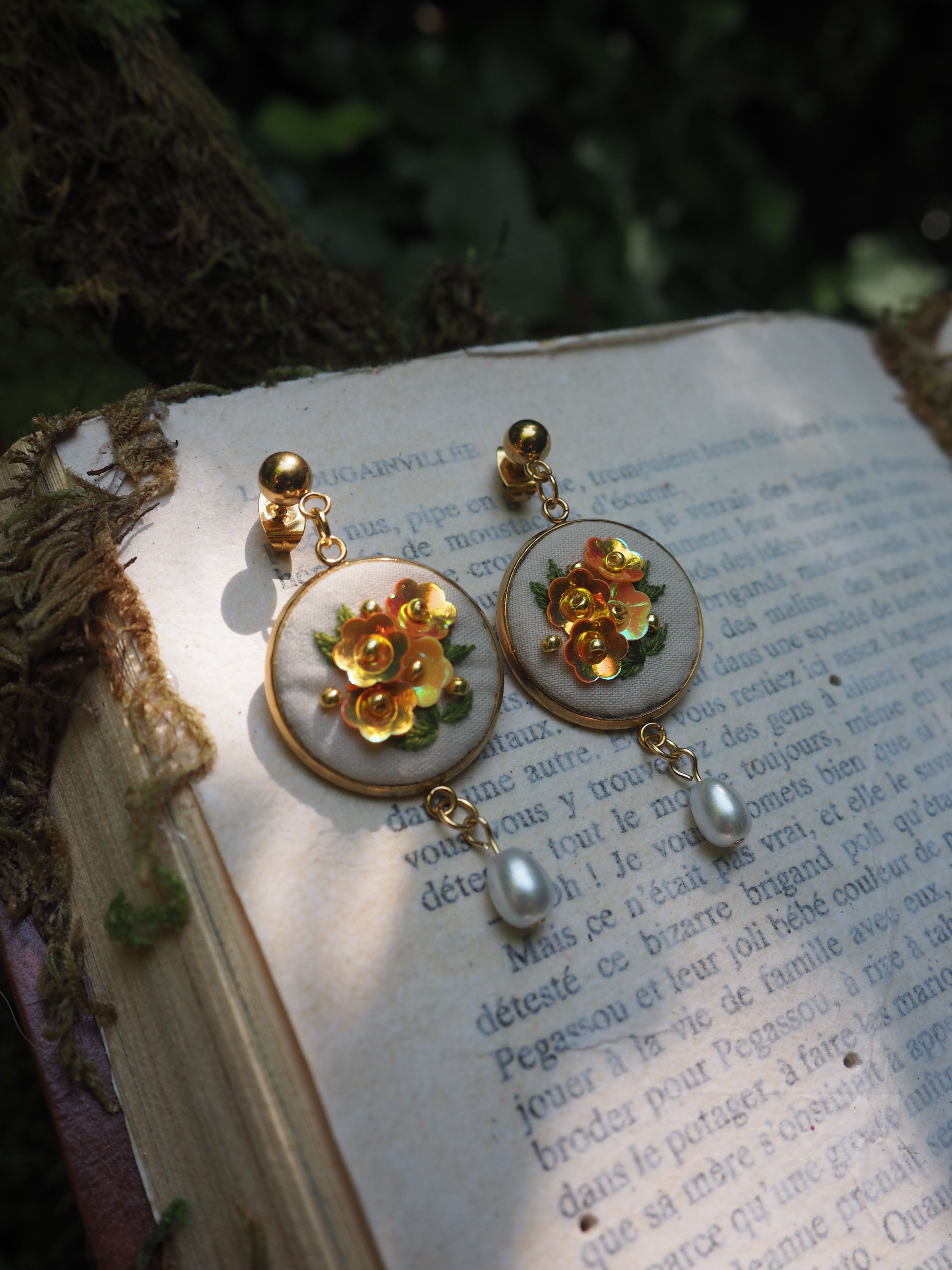 Cette image montre une paire de boucles d'oreilles élégantes posées sur les pages d'un livre en français. Les boucles d'oreilles sont de style vintage ou romantique, composées de médaillons ronds en nacre ou porcelaine blanche, ornés de fleurs dorées en leur centre. Chaque boucle d'oreille est agrémentée d'une petite perle qui pend en dessous. La photo est prise dans un éclairage naturel doux, créant une ambiance poétique et romantique. L'arrière-plan montre du texte français imprimé sur les pages du livre, ajoutant une touche littéraire et française à la composition. L'ensemble crée une atmosphère très délicate et féminine.