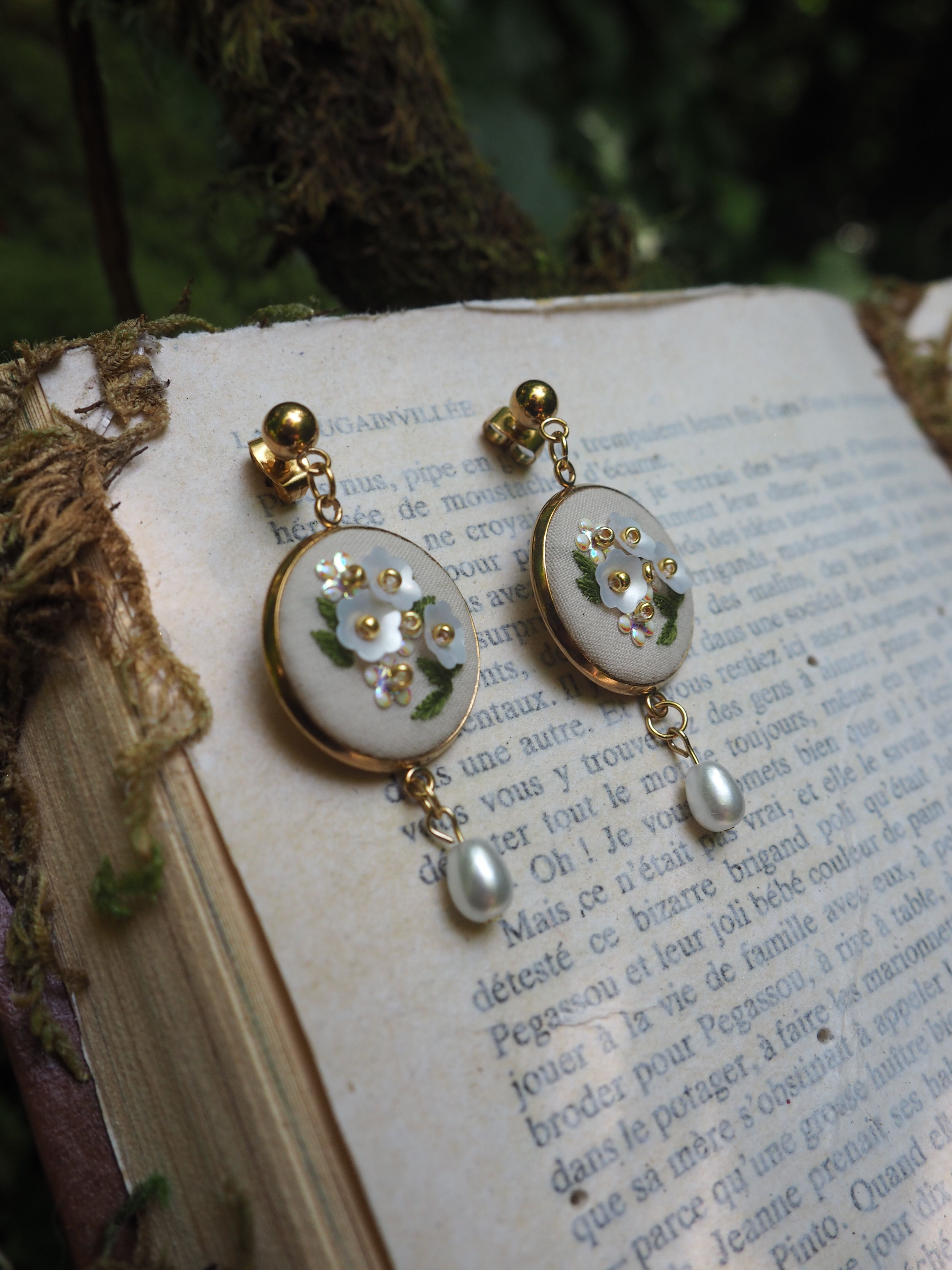 Cette image montre une paire de boucles d'oreilles élégantes posées sur les pages d'un vieux livre en français. Les boucles d'oreilles sont de style vintage avec des disques blancs ornés de petites fleurs dorées et de cristaux. Chaque boucle d'oreille se termine par une perle nacrée suspendue. Le fond montre les pages jaunies du livre avec du texte français visible, et on aperçoit ce qui semble être de l'écorce d'arbre ou du bois naturel dans le coin supérieur de l'image. La composition a un aspect très romantique et poétique, mélangeant les éléments délicats des bijoux avec la littérature classique.