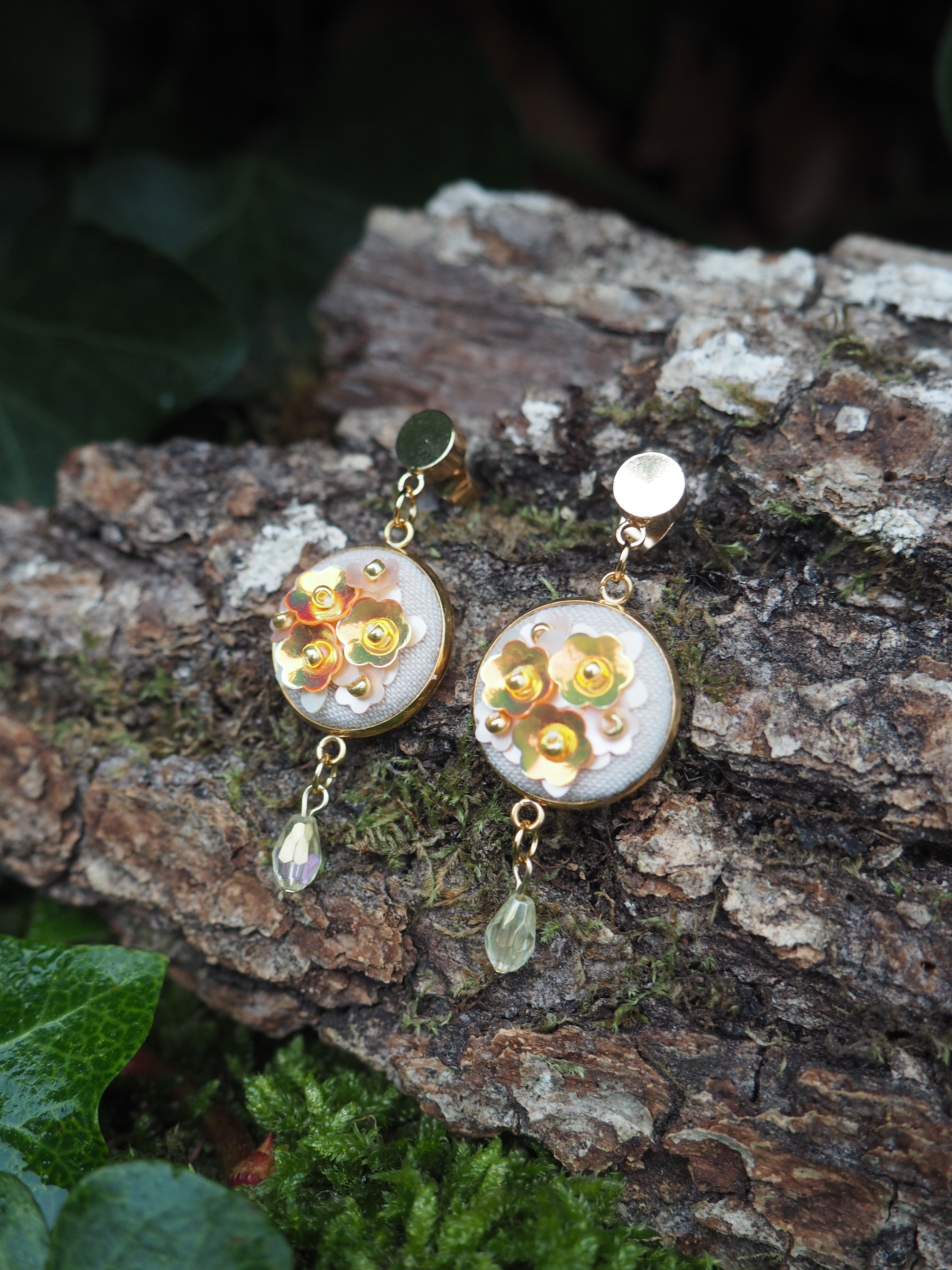 This close-up shot features a pair of elegant, handcrafted earrings resting on a textured tree bark surface, adorned with patches of green moss and hints of dark green foliage in the background. The earrings are designed with a circular, flat base, approximately 2-3 cm in diameter, framed by a thin gold-colored metal rim. The center of each circle is filled with a light, possibly off-white or very pale grey, fabric or material. Adorning this base are several small, shiny, gold-colored sequins shaped like flowers, arranged in a cluster. Some tiny, lighter-colored beads or dots are interspersed among the sequin flowers, adding to the intricate detail. Each earring dangles from a gold-colored stud or clip-on mechanism, connected by a short chain. Below the main circular element, a single, teardrop-shaped bead hangs from another small chain. The bead on the left earring appears to be a clear or iridescent light amber color, while the bead on the right earring is a translucent pale green. The lighting is soft, highlighting the metallic sheen of the gold elements and the sequins, and casting subtle shadows that emphasize the texture of the bark. The overall impression is one of delicate craftsmanship and natural beauty.
