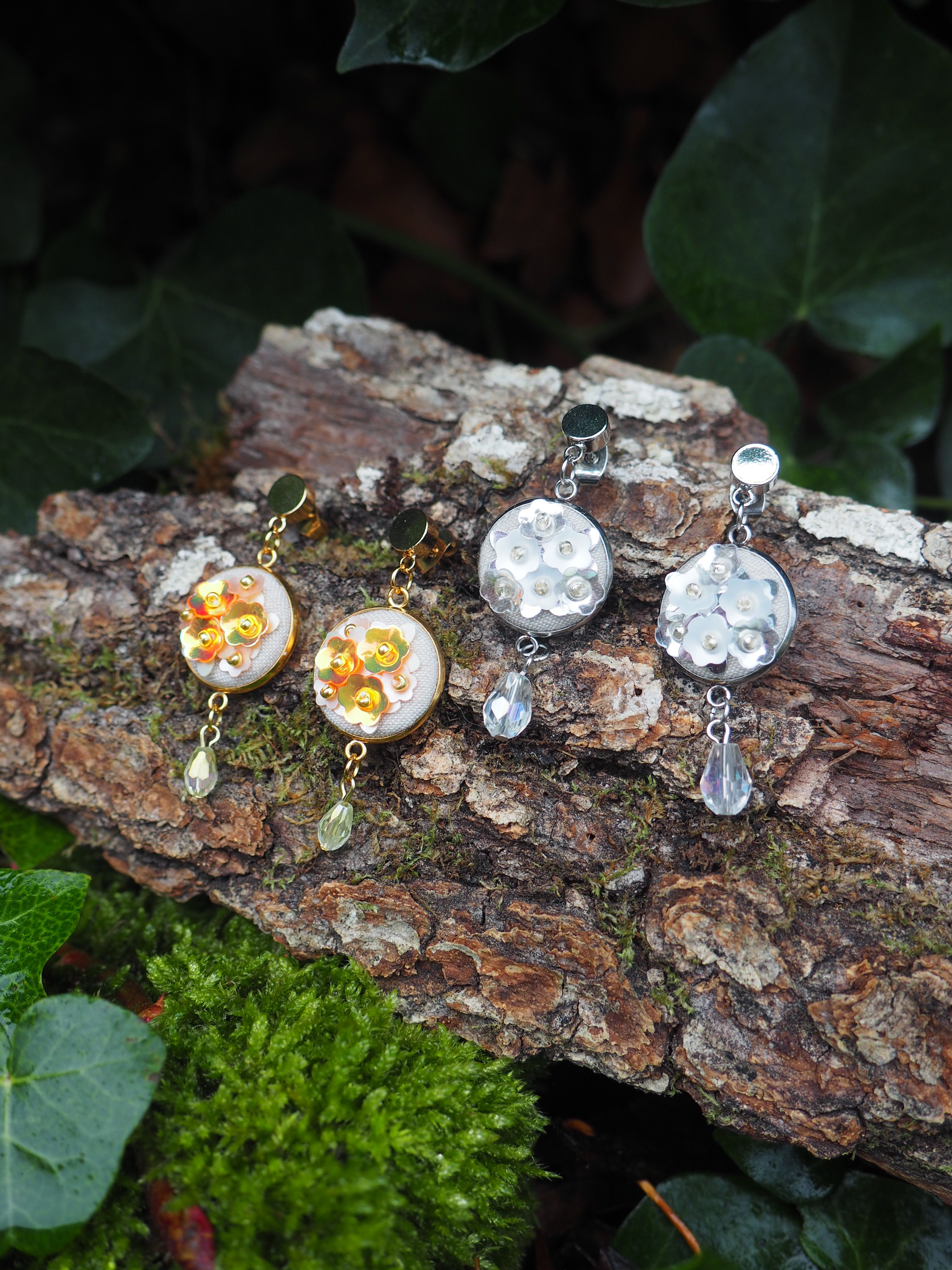 This image features two pairs of decorative earrings, each with a distinct color scheme, resting on a piece of bark. The setting suggests a natural, possibly outdoor, environment with moss and ivy visible. Here's a breakdown of the visual properties: **Overall Composition:**
*   The earrings are the central focus, arranged horizontally across the middle of the frame.
*   They are placed on a textured piece of tree bark, which provides a rustic and organic backdrop.
*   The foreground shows vibrant green moss, adding a pop of color and natural texture.
*   The background is blurred, featuring dark green ivy leaves, which helps to isolate the main subjects.
*   The lighting appears soft and natural, highlighting the details of the earrings and the textures of the bark and moss. **Left Pair of Earrings (Gold/Orange):**
*   These earrings have a gold-toned metal base.
*   The main circular disc of each earring is white or off-white, possibly made of resin or a similar material.
*   Adorning the white disc are numerous small, shiny, golden or orange-colored sequins or beads, arranged in a floral or scattered pattern. They reflect light, giving them a shimmering appearance.
*   Dangling from each earring is a teardrop-shaped bead, which appears to be a light green or yellowish-clear crystal, catching the light. **Right Pair of Earrings (Silver/White):**
*   These earrings have a silver-toned metal base.
*   Similar to the left pair, the main circular disc of each earring is white or off-white.
*   These discs are decorated with small, white, flower-shaped sequins or beads, creating a delicate, textured pattern.
*   Each earring also features a dangling teardrop-shaped bead, which is clear and faceted, resembling a crystal or glass bead, reflecting light brightly. **Background Elements:**
*   **Bark:** The bark is rough and textured, with deep crevices and varying shades of brown, suggesting an aged piece of wood. There are hints of green moss growing on the bark itself.
*   **Moss:** In the foreground, there's a lush patch of bright green moss, providing a soft, velvety contrast to the rough bark.
*   **Ivy:** The blurred background shows dark green ivy leaves, adding depth and a natural, slightly wild feel to the scene. In summary, the image showcases two pairs of handcrafted-looking earrings, one with warm gold and orange tones, and the other with cool silver and white tones, presented in a natural, earthy setting that enhances their aesthetic appeal.