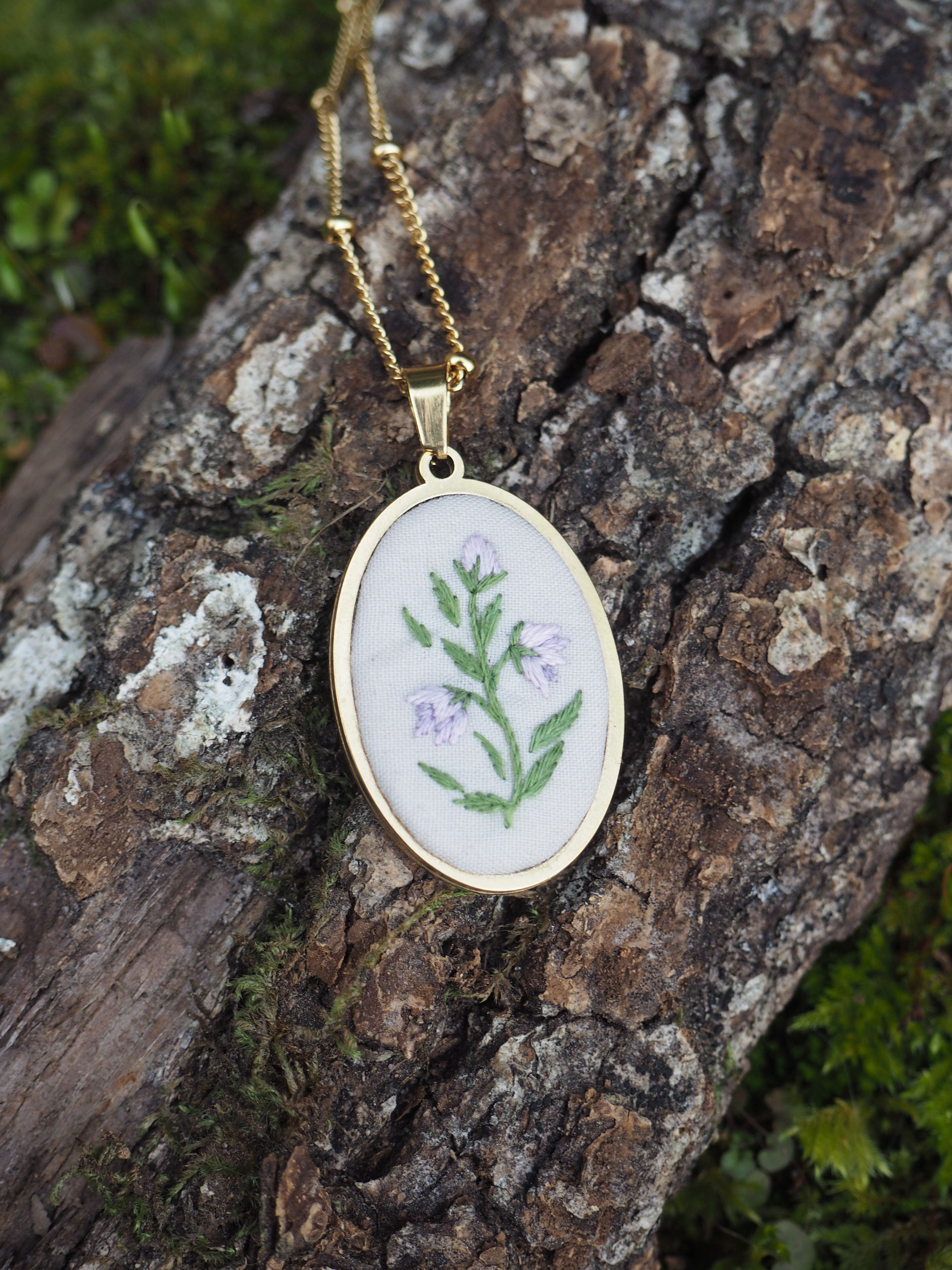 Cette image montre un délicat pendentif de broderie placé sur un morceau d'écorce d'arbre texturée et moussue. Le pendentif est rond, serti dans un cadre doré, et présente une broderie sur fond blanc avec un motif floral simple - des feuilles vertes et de petites fleurs violettes. Il est suspendu à une fine chaîne dorée. Le contraste entre la délicatesse du bijou artisanal et la texture rugueuse de l'écorce naturelle crée une belle composition photographique.