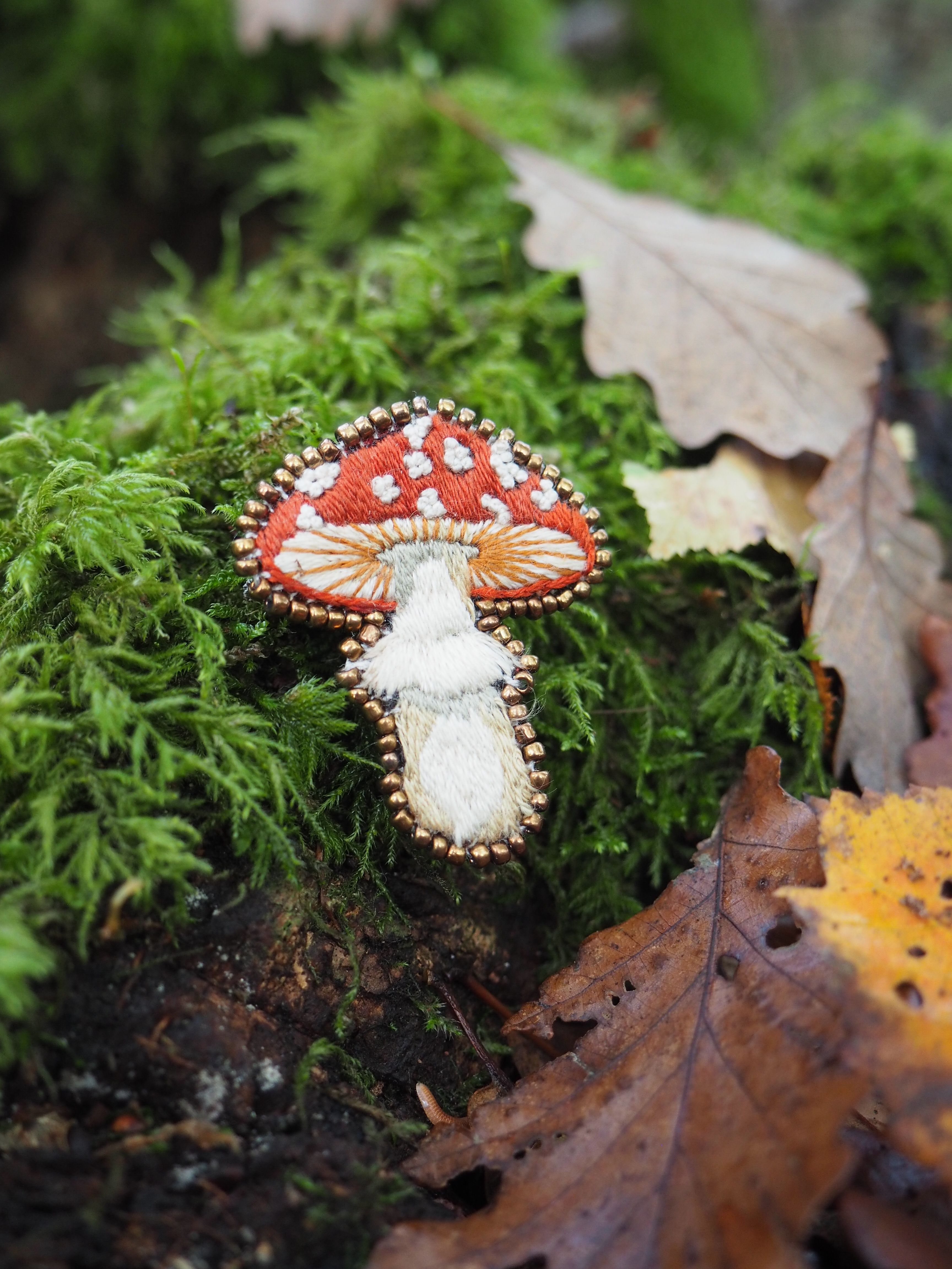 Cette image présente une scène charmante de la nature avec un élément décoratif artistique. Au premier plan, on peut voir un champignon décoratif minutieusement brodé ou cousu, représentant un champignon rouge à pois blancs (rappelant l'amanite tue-mouches) avec un pied blanc. Cette pièce artisanale est délicatement posée sur un lit de mousse verte luxuriante. L'environnement naturel qui l'entoure est composé de :
- Mousse épaisse et verdoyante qui tapisse le sol
- Feuilles mortes aux teintes automnales (brunes et dorées) éparpillées
- Une végétation forestière dense et verte en arrière-plan
- L'ensemble créant une atmosphère de sous-bois humide et paisible L'effet artistique est renforcé par une profondeur de champ qui maintient le champignon brodé net au premier plan tandis que l'arrière-plan reste légèrement flou, créant un contraste saisissant entre l'art textile et la nature authentique. Cette composition évoque un conte de fées ou un monde fantastique où l'artisanat se mêle harmonieusement à l'environnement forestier.