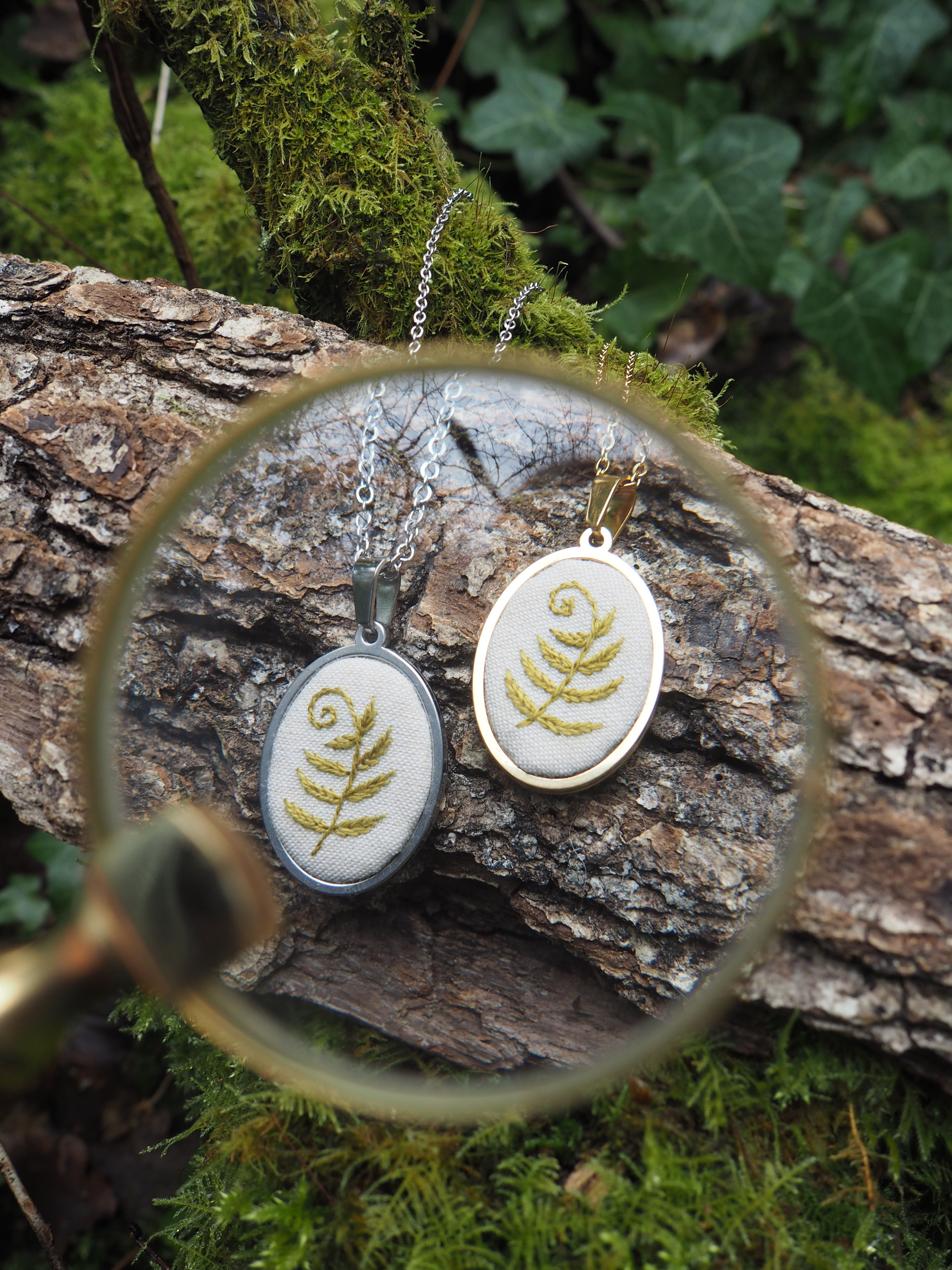 Cette image présente une composition artistique et naturelle, centrée sur deux pendentifs ovales, chacun orné d'un motif de fougère stylisé. Au premier plan, une loupe dorée est positionnée de manière à encadrer partiellement les pendentifs, créant un effet de mise au point et d'agrandissement sur les détails des bijoux. La monture dorée de la loupe est visible sur le côté gauche et en bas de l'image. Les deux pendentifs sont suspendus à des chaînes fines et reposent sur un tronc d'arbre horizontal, recouvert de mousse verte et de lichens, suggérant un environnement forestier ou naturel.
Le pendentif de gauche a un cadre argenté et un fond clair (peut-être blanc cassé ou crème) sur lequel est brodé ou dessiné un motif de fougère en spirale, de couleur dorée ou vert clair brillant.
Le pendentif de droite a un cadre doré et un fond similaire, avec un motif de fougère identique, également doré ou vert clair brillant. L'arrière-plan est flou, mais on distingue des feuillages verts, probablement des lierres et d'autres plantes forestières, renforçant l'ambiance naturelle et organique de la scène. La lumière semble douce et diffuse, mettant en valeur les textures de la mousse et le brillant des pendentifs. L'ensemble de l'image évoque un sentiment de délicatesse, de nature et d'artisanat, avec une attention particulière portée aux détails des pendentifs et à leur présentation dans un cadre authentique.