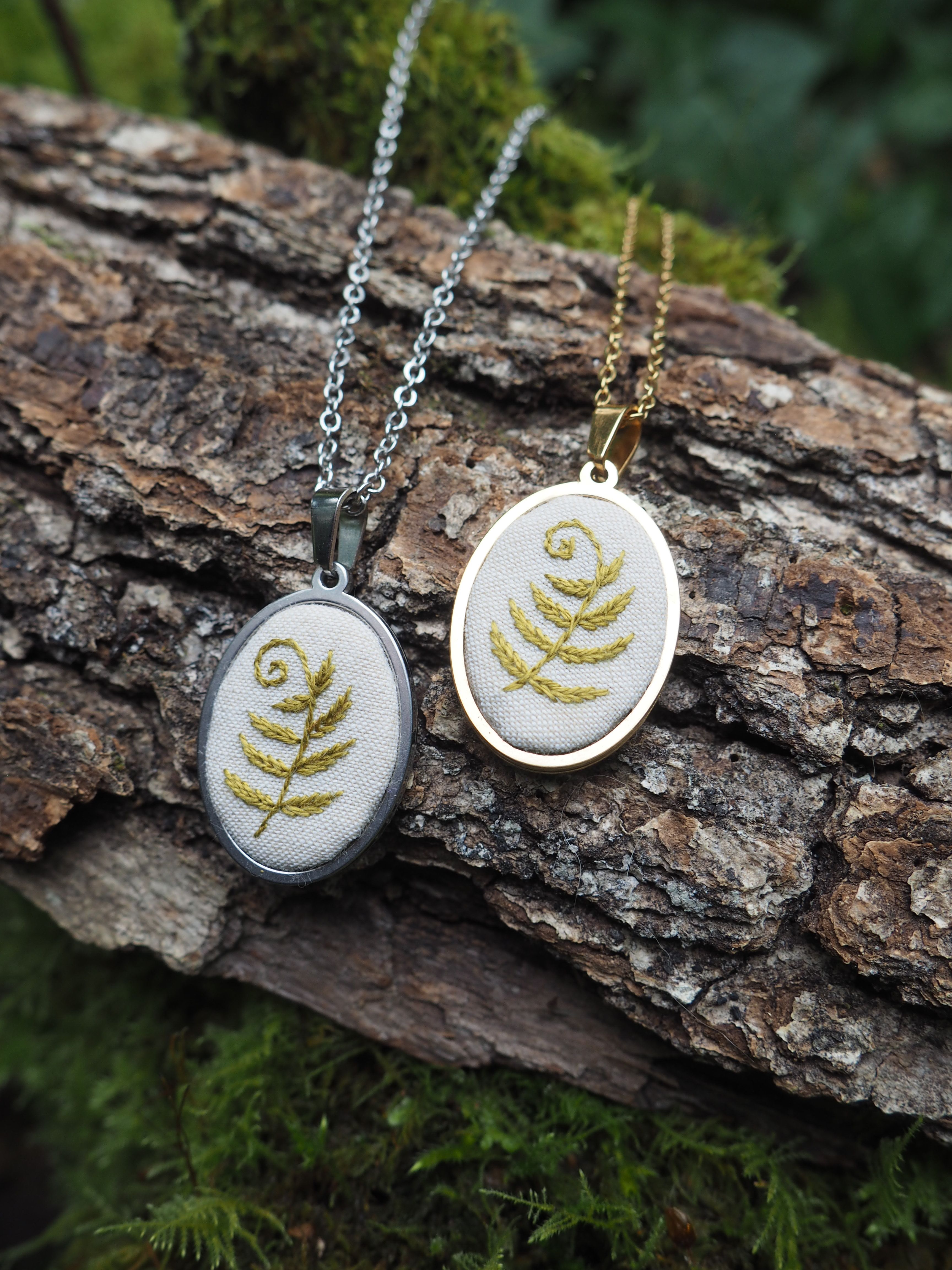 Cette image présente deux pendentifs ovales, chacun orné d'une broderie délicate représentant une fougère stylisée. Les pendentifs sont posés sur un morceau de bois brut, recouvert de mousse par endroits, ce qui crée un contraste intéressant entre la nature et l'artisanat. Voici une description plus détaillée des éléments visuels : *   **Les Pendentifs :**
    *   **Pendentif de gauche :** Il a un contour métallique de couleur argentée ou gris foncé. La broderie à l'intérieur est une fougère de couleur dorée ou jaune pâle, réalisée sur un fond de tissu clair, probablement blanc cassé ou crème. La chaîne attachée à ce pendentif est également de couleur argentée.
    *   **Pendentif de droite :** Il présente un contour métallique de couleur dorée. La broderie de la fougère est également dorée ou jaune pâle, sur un fond de tissu clair similaire à l'autre pendentif. La chaîne de ce pendentif est de couleur dorée.
    *   Les motifs de fougère sont identiques sur les deux pendentifs, avec une forme en spirale caractéristique des jeunes frondes de fougère (crosses de fougère). *   **Le Support (Bois) :**
    *   Les pendentifs reposent sur un morceau de bois, qui semble être une branche ou un tronc d'arbre.
    *   La texture du bois est rugueuse et écorcée, avec des crevasses et des motifs naturels.
    *   Des touches de mousse verte sont visibles sur le bois, ajoutant une touche organique et naturelle à la composition. *   **L'Arrière-plan :**
    *   L'arrière-plan est flou, suggérant une faible profondeur de champ, ce qui met en valeur les pendentifs.
    *   On distingue des teintes de vert foncé, probablement de la végétation ou de la mousse, renforçant l'ambiance naturelle et forestière de la scène. *   **Lumière et Ambiance :**
    *   La lumière semble naturelle et douce, éclairant bien les pendentifs et faisant ressortir les détails des broderies et les reflets des métaux.
    *   L'ambiance générale est calme, naturelle et artisanale, évoquant la beauté de la nature et le travail manuel. En résumé, l'image met en scène deux pendentifs brodés de fougères, présentés de manière esthétique sur un support en bois naturel, soulignant leur caractère artisanal et leur inspiration botanique.