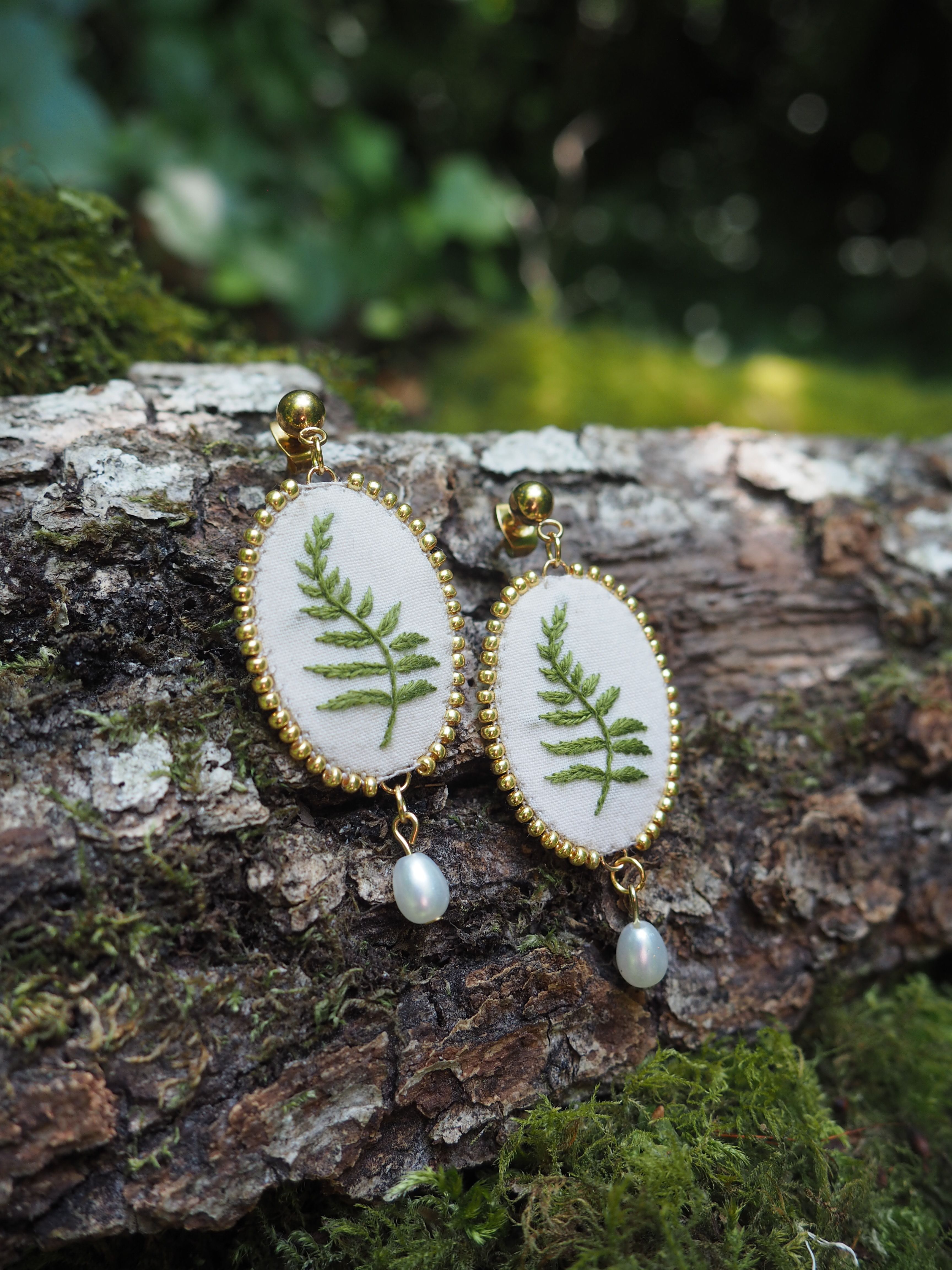 Cette image montre une paire de boucles d'oreilles artisanales très délicates. Chaque boucle d'oreille est composée d'un médaillon ovale avec un cadre doré décoratif. À l'intérieur de chaque médaillon, on peut voir une véritable feuille de fougère pressée sur un fond blanc. Au bas de chaque boucle d'oreille pend une petite perle blanche. Les boucles d'oreilles sont photographiées sur ce qui semble être un morceau d'écorce d'arbre couvert de mousse, créant un cadre naturel qui complimente parfaitement le motif botanique des bijoux. L'arrière-plan est flou mais montre des tons verts naturels, suggérant un environnement forestier. Ce sont des bijoux qui capturent vraiment l'essence de la nature, combinant élégamment des éléments naturels avec un design raffiné.