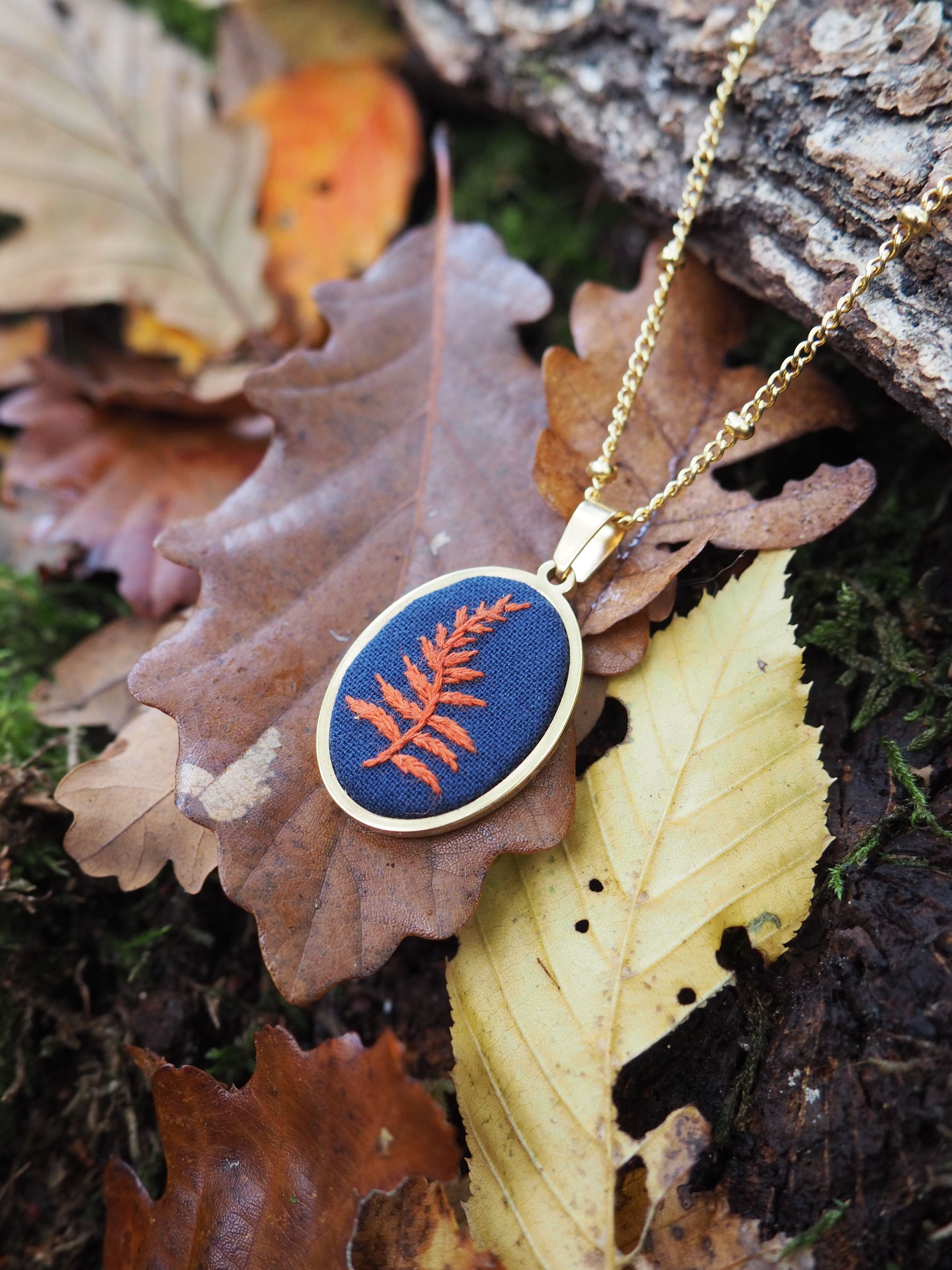Cette image présente un magnifique pendentif artisanal posé sur un tapis de feuilles d'automne. Le bijou se compose d'un médaillon ovale avec un fond bleu profond sur lequel est délicatement brodée ou appliquée une fougère de couleur cuivre/orange. Le pendentif est suspendu à une chaîne dorée fine et élégante. L'ensemble repose sur des feuilles mortes aux teintes automnales variées - du brun doré au jaune pâle en passant par l'orange - créant une harmonie parfaite avec les couleurs du bijou. On distingue également de l'écorce d'arbre et quelques éléments naturels qui renforcent l'atmosphère forestière et saisonnière de la composition. La photographie met en valeur le contraste entre les matières : la finesse du travail artisanal du pendentif et la texture organique des feuilles séchées. L'éclairage naturel fait ressortir les détails de la broderie et donne une ambiance chaleureuse à l'ensemble, évoquant une promenade en forêt à l'automne.