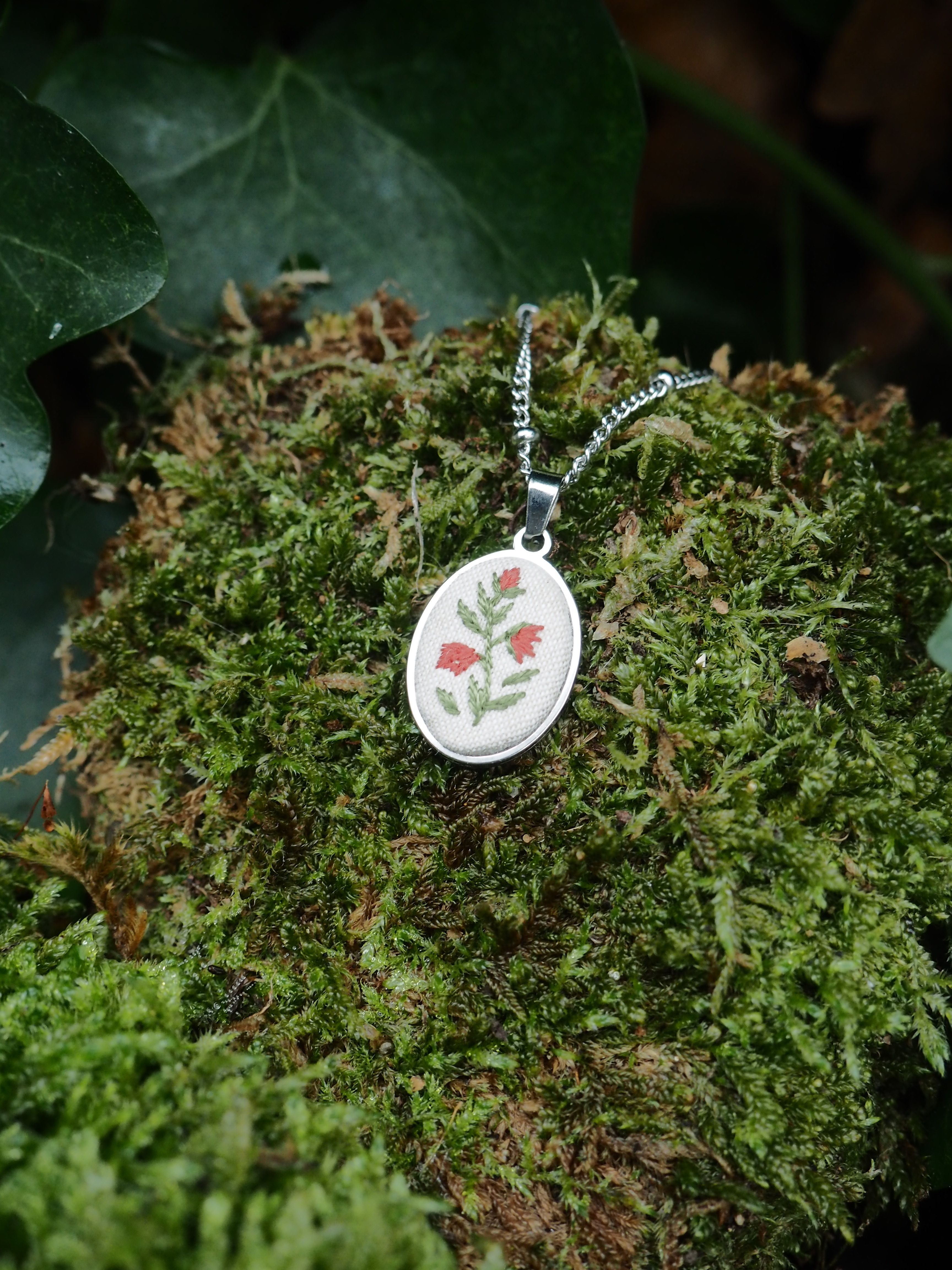 This image features a close-up shot of an oval-shaped pendant with a silver-colored chain, resting on a bed of vibrant green moss. The pendant itself is the focal point, showcasing a delicate embroidery of three red bell-shaped flowers with green stems and leaves, set against a light, possibly white or cream, fabric background. The embroidery is detailed, giving the flowers a slightly textured appearance. The pendant has a smooth, reflective silver-toned bezel surrounding the embroidered insert. The chain attached to the pendant is a fine, silver-colored link chain, visible extending upwards from the pendant. The moss beneath the pendant is rich in texture and varying shades of green, suggesting a natural, outdoor setting. In the background, out of focus, there are hints of darker green foliage, possibly ivy leaves, further emphasizing the natural theme. The lighting appears soft and natural, highlighting the details of the pendant and the moss without harsh shadows. The overall impression is one of handcrafted beauty and a connection to nature.