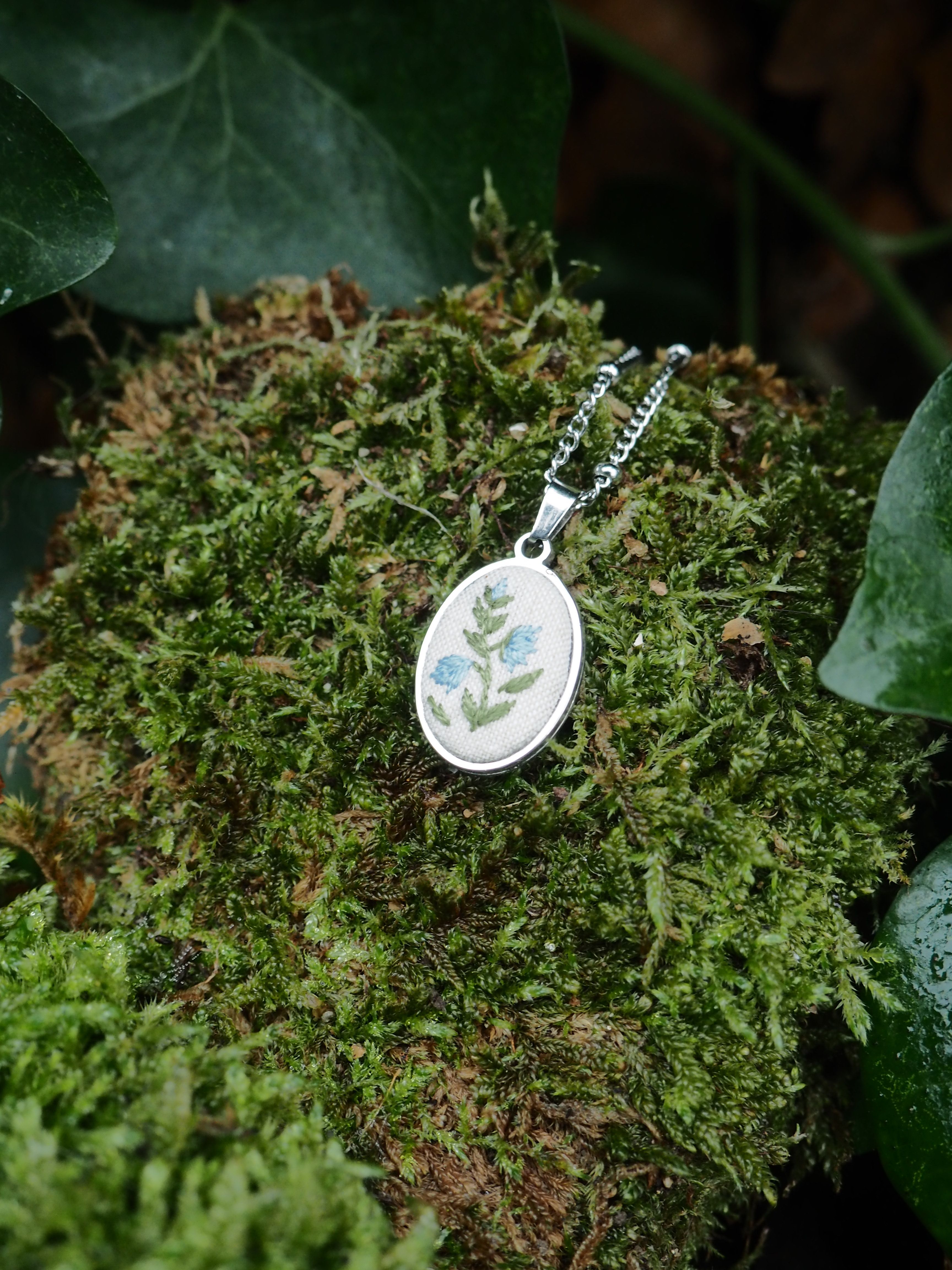 This image features a close-up shot of an oval-shaped pendant with a silver-colored chain, resting on a bed of vibrant green moss. The pendant itself has a white or light-colored background with an embroidered or painted design of light blue flowers with green stems and leaves. The design appears delicate and natural. The moss provides a textured, organic backdrop, with varying shades of green and some darker, possibly brown, undertones suggesting depth and natural growth. In the upper left and lower right corners, out-of-focus dark green leaves, possibly ivy, frame the scene, adding to the natural and earthy aesthetic. The lighting highlights the pendant and the moss, creating a soft, natural feel.