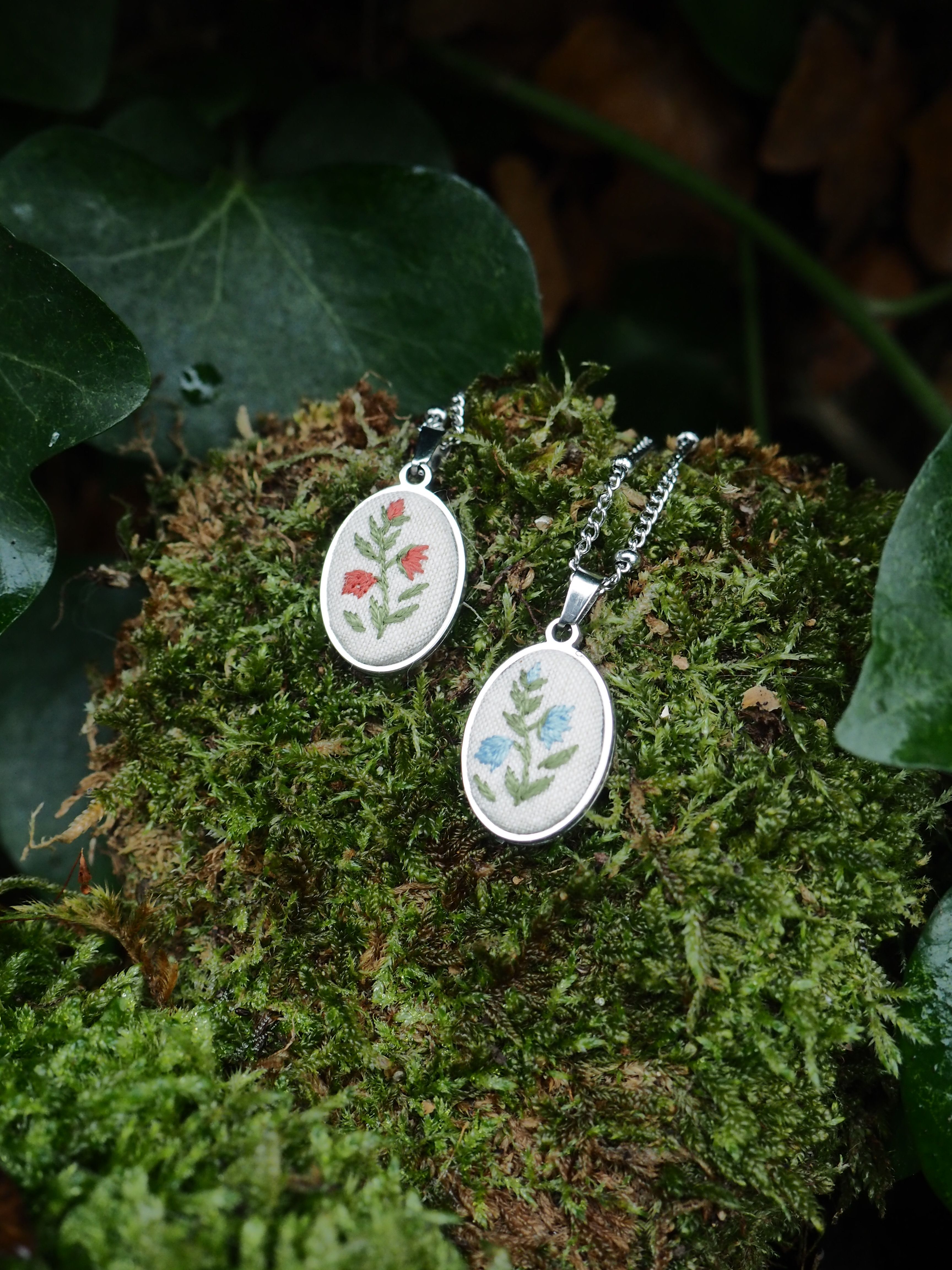 This image features two oval-shaped pendants, each with a delicate embroidered floral design, resting on a bed of vibrant green moss. The pendants are silver-toned and have a white fabric base for the embroidery. The pendant on the left displays a design of two red flowers with green stems and leaves. The pendant on the right showcases a design of two blue flowers with green stems and leaves. Both pendants are attached to silver-colored chains, which are partially visible. The moss provides a natural and textured backdrop, with varying shades of green and some brown undertones, suggesting a forest or garden setting. In the background, out of focus, are dark green leaves, possibly ivy, further enhancing the natural aesthetic of the scene. The lighting appears soft, highlighting the details of the pendants and the texture of the moss.