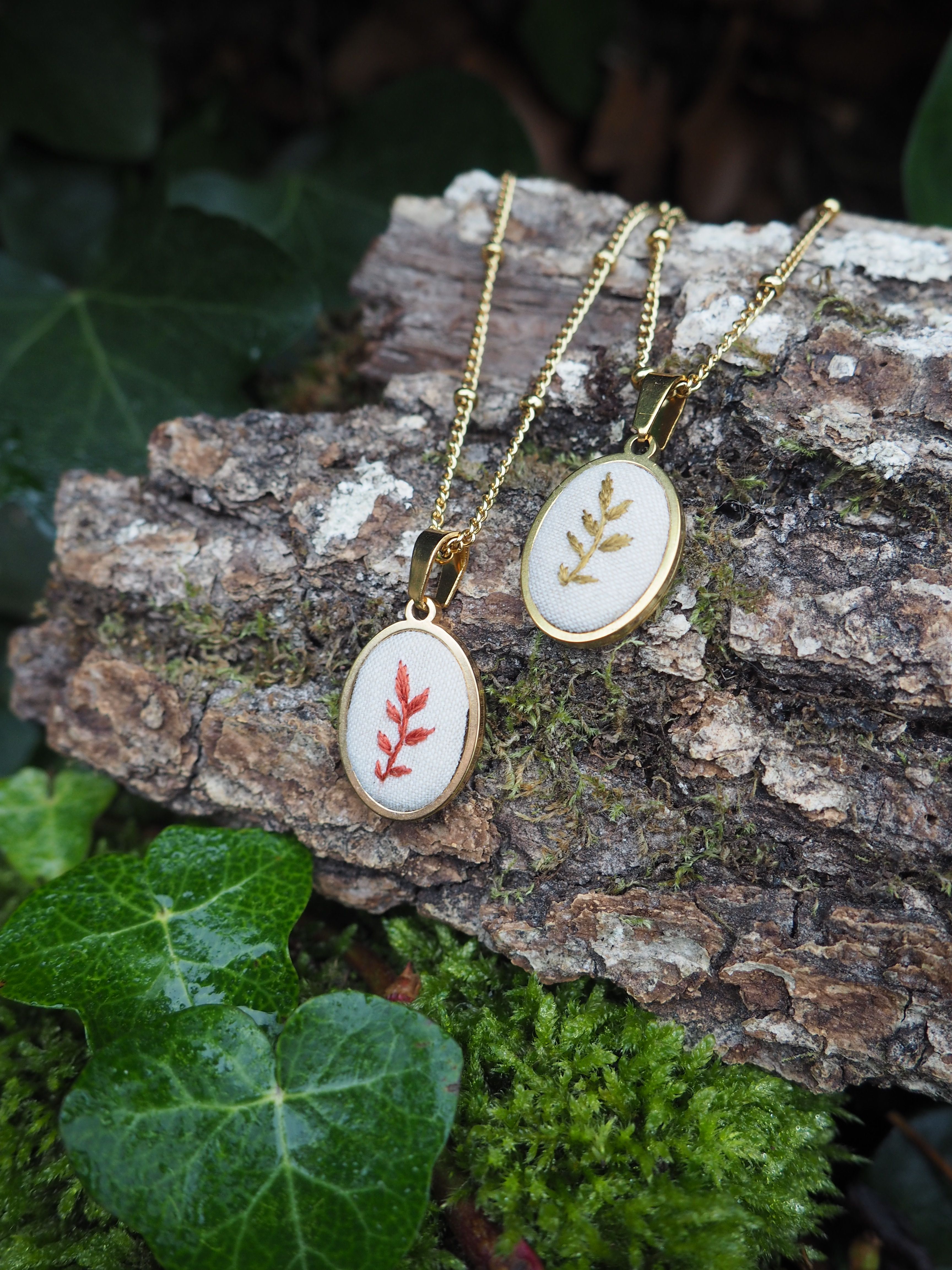 This image features two delicate oval-shaped pendants with gold-colored chains, resting on a piece of textured tree bark. The pendants are identical in shape and size, each featuring a white fabric insert with an embroidered leaf design. The pendant on the left displays a reddish-brown leaf design, while the one on the right has a golden-yellow leaf design. Both pendants are framed by a shiny gold-colored bezel. The chains are thin and have small, evenly spaced beads along their length. The tree bark beneath the pendants is rough and dark brown, with visible grooves and some patches of green moss. To the left and bottom of the pendants, there are vibrant green ivy leaves, some of which appear to have water droplets on them, suggesting a damp or natural outdoor setting. The background is softly blurred, showing hints of darker foliage. The overall impression is one of natural beauty and handcrafted elegance, with the jewelry harmonizing with its organic surroundings.