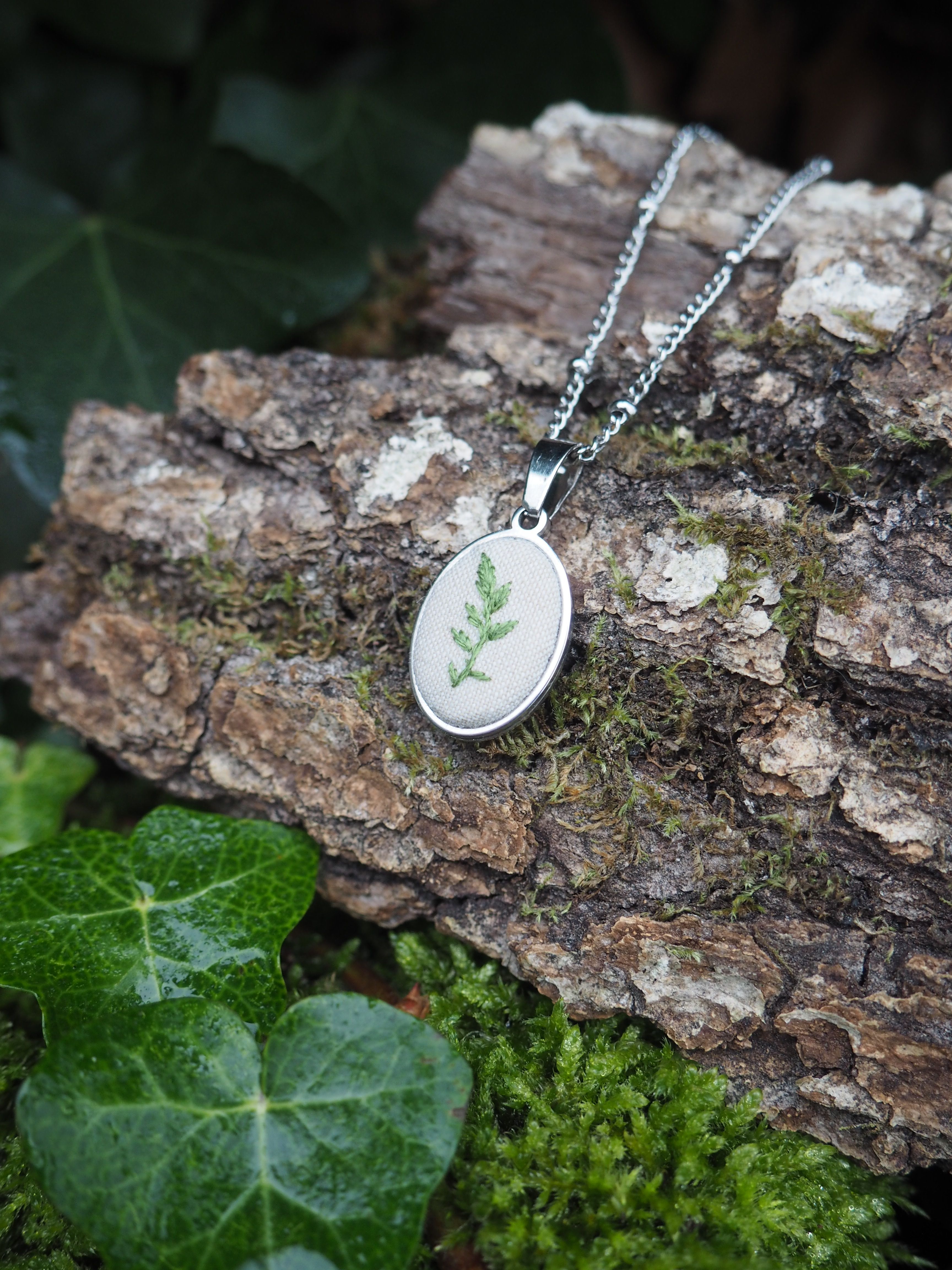 This image features a close-up, eye-level shot of a silver pendant necklace resting on a piece of tree bark. The pendant is oval-shaped with a silver frame and a white fabric insert. Embroidered on the white fabric is a delicate green fern leaf design. The silver chain of the necklace extends upwards and to the right, partially visible. The tree bark, which serves as the primary surface for the necklace, is rough and textured, displaying shades of brown and grey. Patches of vibrant green moss are visible on and around the bark, adding to the natural aesthetic. In the foreground, to the left, several glossy green ivy leaves are visible, some with small water droplets on their surface, suggesting a damp or recently wet environment. The background is softly blurred, showing more dark green foliage, likely more ivy or other plants, contributing to a natural, earthy setting. The lighting appears soft and natural, highlighting the details of the pendant and the textures of the surrounding elements. The overall impression is one of natural beauty and handcrafted elegance.