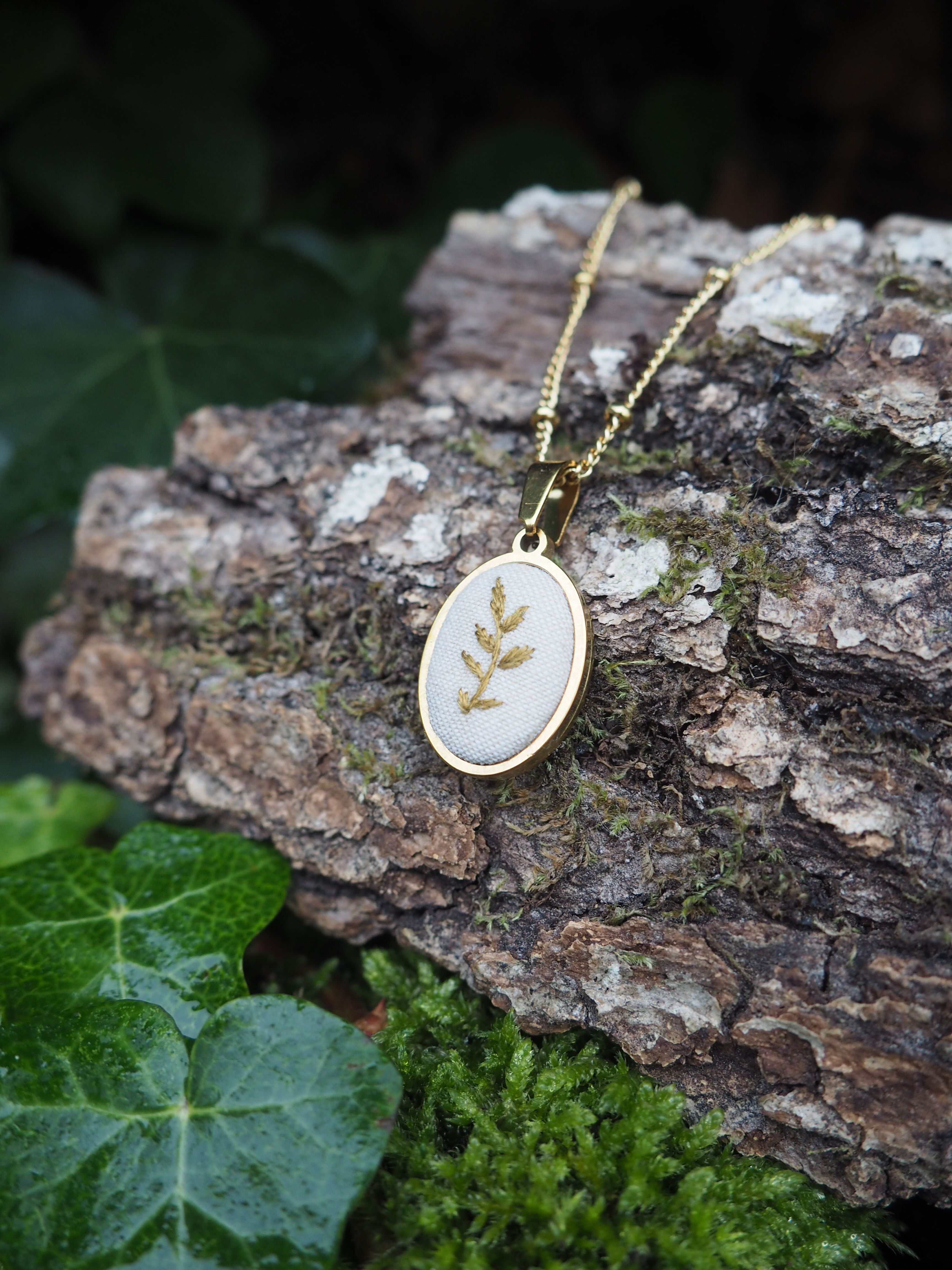 This close-up shot features a delicate gold-colored necklace with an oval pendant, resting on a piece of textured tree bark. The pendant is the focal point, showcasing a light, possibly white or cream-colored, fabric or enamel base. Embroidered or depicted on this base is a stylized golden leaf or sprig design, adding a touch of nature-inspired elegance. The pendant is framed by a thin gold-colored bezel. The necklace chain is also gold-colored and appears to be a fine, beaded or delicate link chain, partially visible as it drapes across the bark. The tree bark provides a rustic and natural backdrop, with its rough texture and varying shades of brown and grey. Patches of vibrant green moss are visible on the bark, enhancing the organic feel of the scene. In the foreground and to the left, lush green ivy leaves are visible, some with a slight sheen, suggesting they might be damp or recently watered. The background is softly blurred, with hints of dark green foliage, further emphasizing the natural setting. The lighting appears soft and natural, highlighting the metallic gleam of the necklace and the intricate details of the pendant, while creating a gentle contrast with the earthy tones of the bark and foliage.