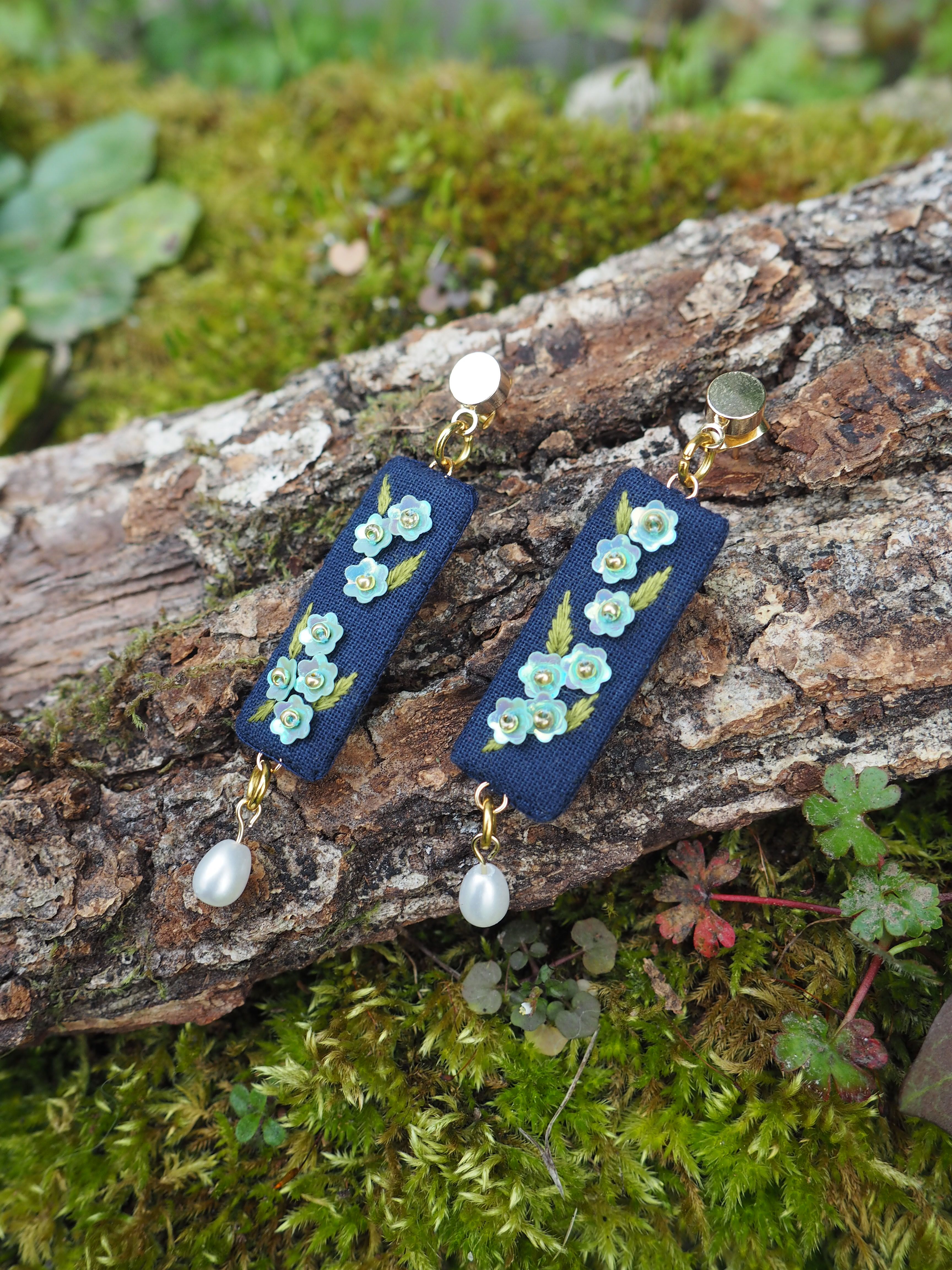 Cette image montre une paire de boucles d'oreilles artisanales posées sur un tronc d'arbre couvert de mousse. Les boucles d'oreilles sont composées d'une base en tissu ou cuir bleu marine, décorée de petites fleurs turquoise ou vert d'eau. Au bas de chaque boucle d'oreille pend une perle blanche nacrée. Les bijoux sont photographiés dans un cadre naturel, avec de la mousse verte et des petites plantes en arrière-plan, ce qui crée une ambiance naturelle et romantique. Le contraste entre le bleu des boucles d'oreilles et le vert de la mousse met particulièrement en valeur ces bijoux délicats.
