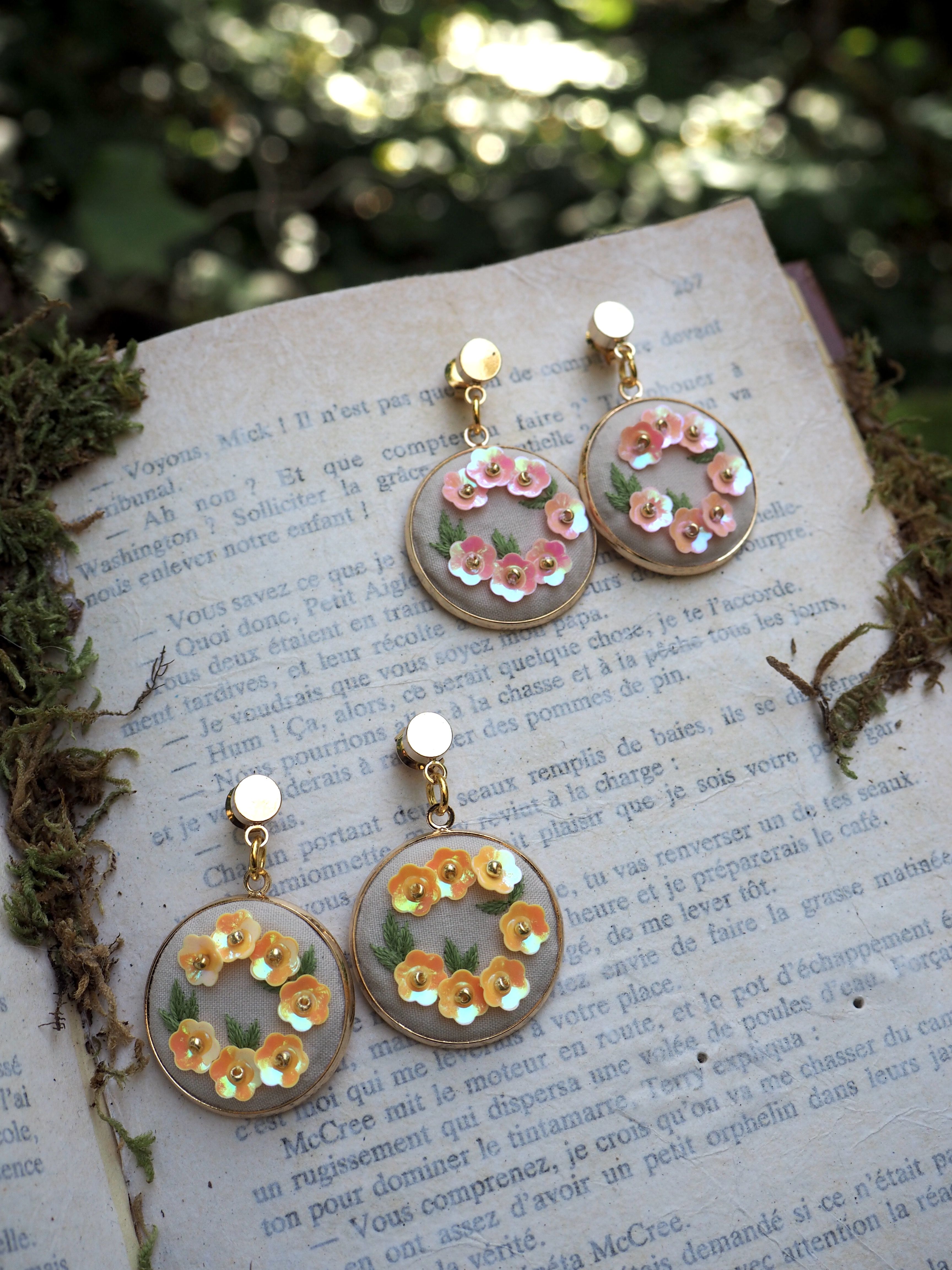 This image shows two pairs of delicate handmade earrings displayed on what appears to be an old French text or book page. The earrings feature a circular design with small decorative elements arranged in a ring pattern. One pair has pink/coral colored details while the other pair has yellow/gold colored accents. The earrings appear to be made with clear resin or acrylic bases with clip-on or post backings. They're photographed in natural lighting with some moss or greenery visible around the edges of the book page, creating a whimsical, vintage-inspired aesthetic. The overall composition has a romantic, artisanal quality that suggests these are handcrafted jewelry pieces.