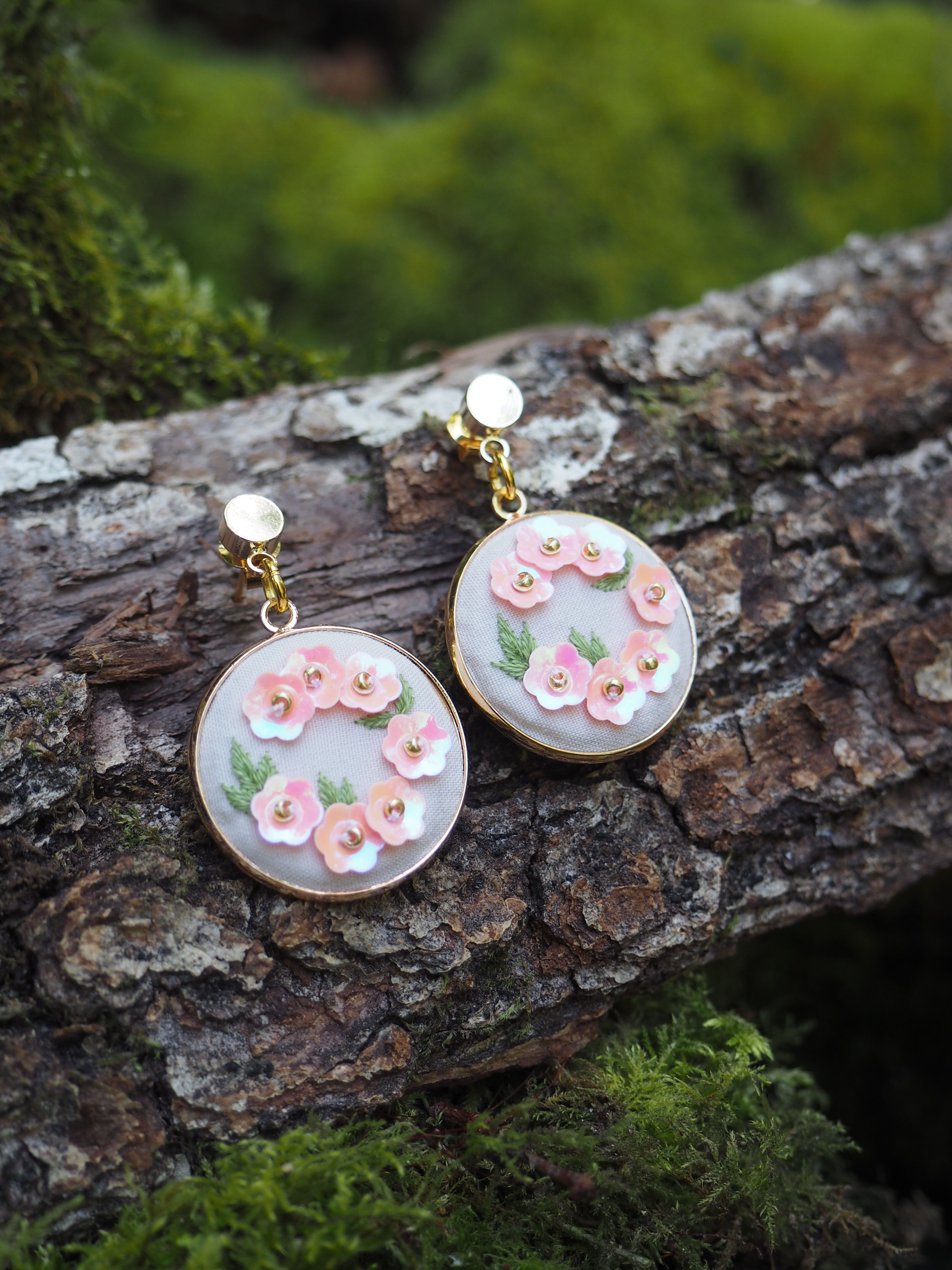 Cette image montre une paire de boucles d'oreilles artisanales délicates posées sur un morceau d'écorce d'arbre moussu. Les boucles d'oreilles sont de forme circulaire avec un fond blanc ou nacré, décorées de petites fleurs roses en relief et de feuilles vertes. Elles semblent être faites à la main, probablement en polymère ou en résine, avec des attaches dorées. La composition est très naturelle et romantique, avec le contraste entre la texture rugueuse de l'écorce et la délicatesse des bijoux. La mousse verte en arrière-plan ajoute une touche naturelle et féerique à la photo.