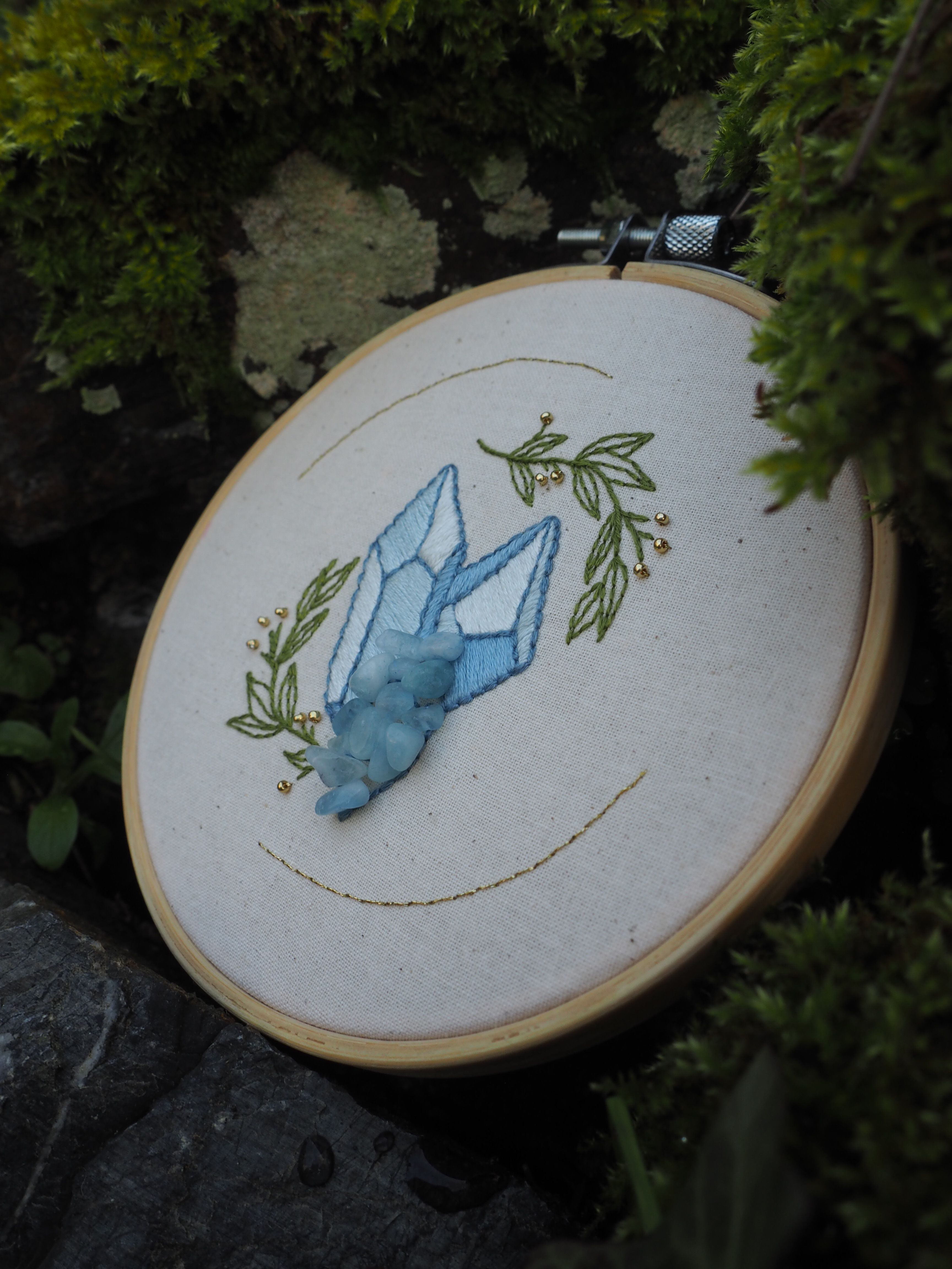 Cette image montre un ouvrage de broderie dans un cercle à broder traditionnel en bois. La broderie représente un cristal ou une pierre précieuse de couleur bleu clair, entourée d'éléments végétaux brodés en vert olive. Le motif est réalisé sur un tissu blanc, probablement de la toile à broder. La photo est prise dans un environnement extérieur, avec de la verdure floue en arrière-plan, ce qui crée une belle mise en scène naturelle pour cette pièce d'artisanat. Le travail de broderie semble délicat et soigné, avec des points qui créent un bel effet d'ombrage sur le cristal.