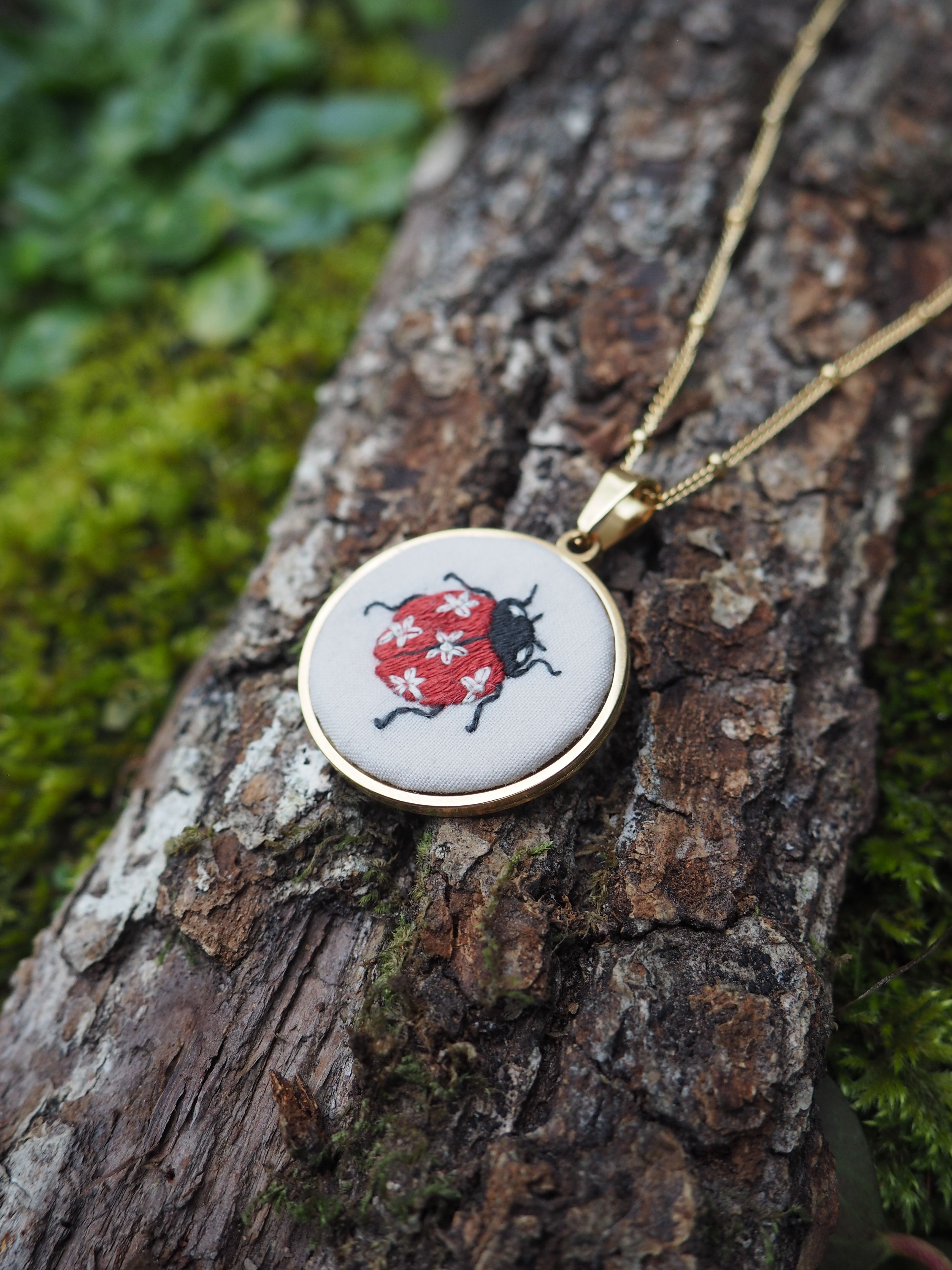 Cette image montre un pendentif artisanal très délicat. Il s'agit d'une broderie miniature encadrée dans un support rond doré, porté en collier sur une chaîne dorée. Le motif brodé représente un scarabée ou un insecte noir sur un fond rouge. Le pendentif est photographié posé sur un tronc d'arbre recouvert de mousse verte, ce qui crée un beau contraste naturel avec le bijou. L'image a une ambiance très nature et artisanale, mettant en valeur ce bijou unique fait main.