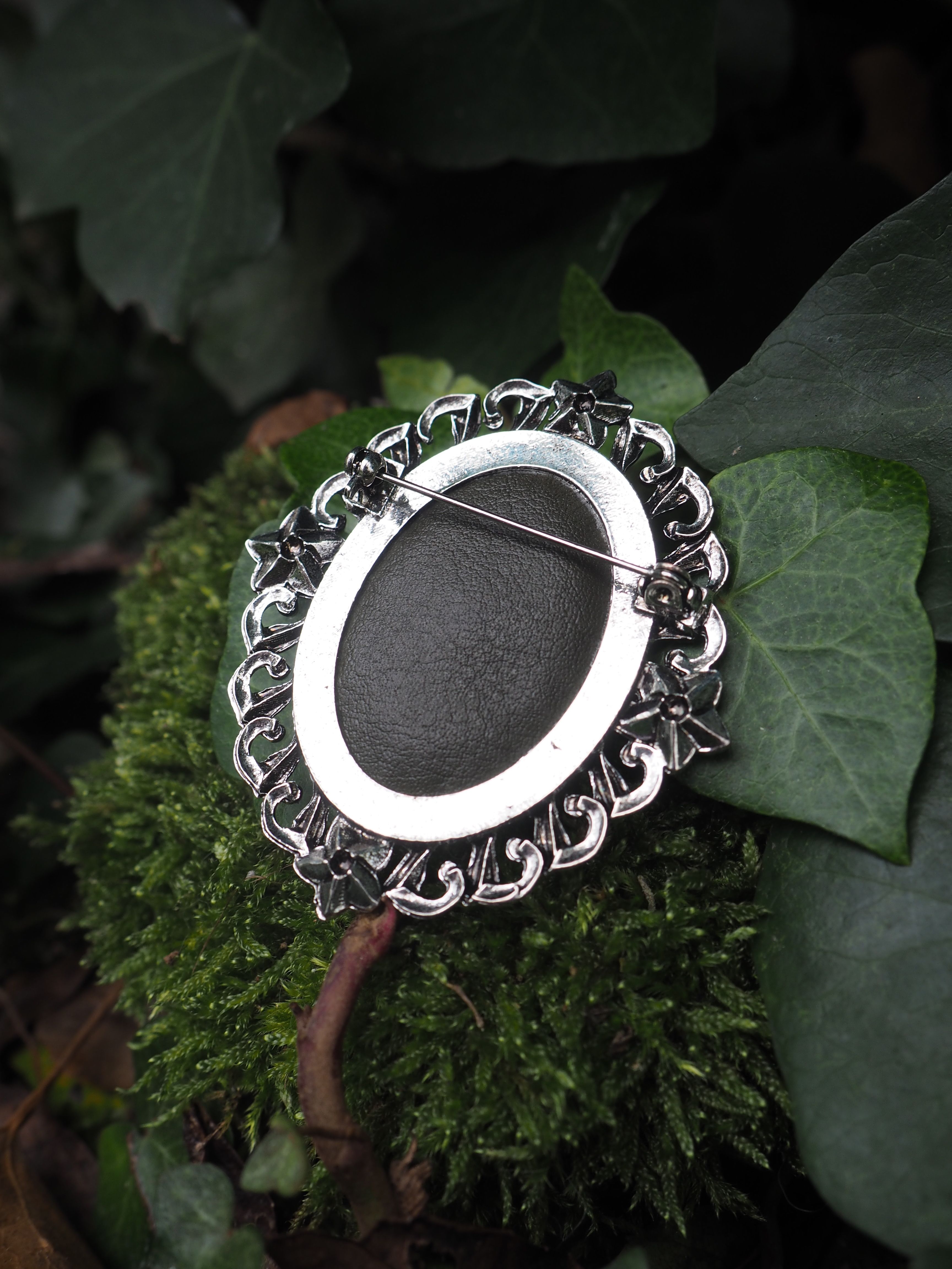 Cette image présente une broche ou un pendentif de style vintage posé délicatement sur des feuilles vertes dans un environnement naturel.  **Caractéristiques du bijou :**
- Forme ovale avec un centre noir mat (probablement en onyx ou une pierre similaire)
- Monture en métal argenté (vraisemblablement de l'argent) avec des motifs décoratifs ciselés
- Bordure ornementée de volutes et de motifs floraux en relief
- Style rétro ou victorien avec un travail artisanal détaillé **Mise en scène :**
- Le bijou repose sur une feuille verte texturée, possiblement une feuille de géranium ou de plante similaire
- L'arrière-plan montre d'autres feuillages dans des tons verts variés
- L'éclairage naturel met en valeur les contrastes entre le métal brillant, la pierre noire et la verdure environnante
- La composition crée une atmosphère élégante et naturelle, soulignant la beauté intemporelle du bijou Cette photographie met particulièrement bien en valeur le contraste entre l'objet manufacturé raffiné et son écrin végétal naturel.