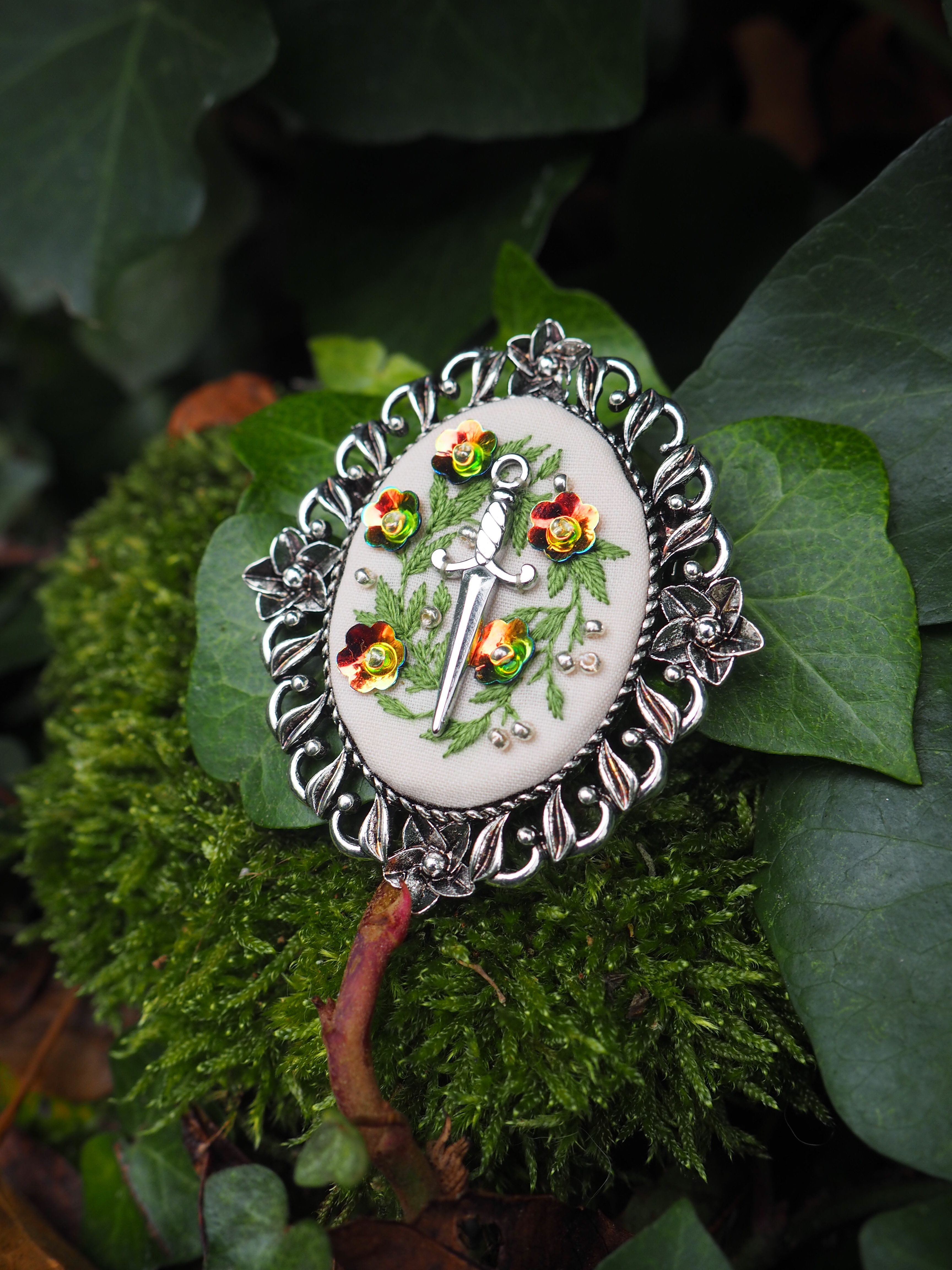 Cette image présente un magnifique bijou artisanal posé sur un lit de mousse et de feuillage naturel. Il s'agit d'une broche ou d'un pendentif de forme circulaire avec un design élaboré : **Caractéristiques du bijou :**
- **Monture** : Un cadre en métal argenté travaillé avec des motifs décoratifs en relief tout autour du pourtour
- **Centre** : Un médaillon en porcelaine ou céramique blanche ornée d'un motif peint à la main représentant des fleurs et du feuillage dans des tons verts
- **Détails colorés** : De petites pierres ou cabochons de couleur ambrée/dorée sont sertis dans la monture métallique
- **Style** : Le bijou évoque l'artisanat traditionnel ou vintage, avec un travail minutieux et raffiné **Mise en scène :**
L'objet est photographié dans un environnement naturel sur de la mousse verte et des feuilles, créant un contraste harmonieux entre l'artifice du bijou et la nature environnante. L'éclairage met en valeur les détails du travail métallique et fait ressortir les couleurs délicates du motif central. Ce type de bijou semble être une pièce d'artisanat de qualité, possiblement inspirée de styles européens traditionnels.