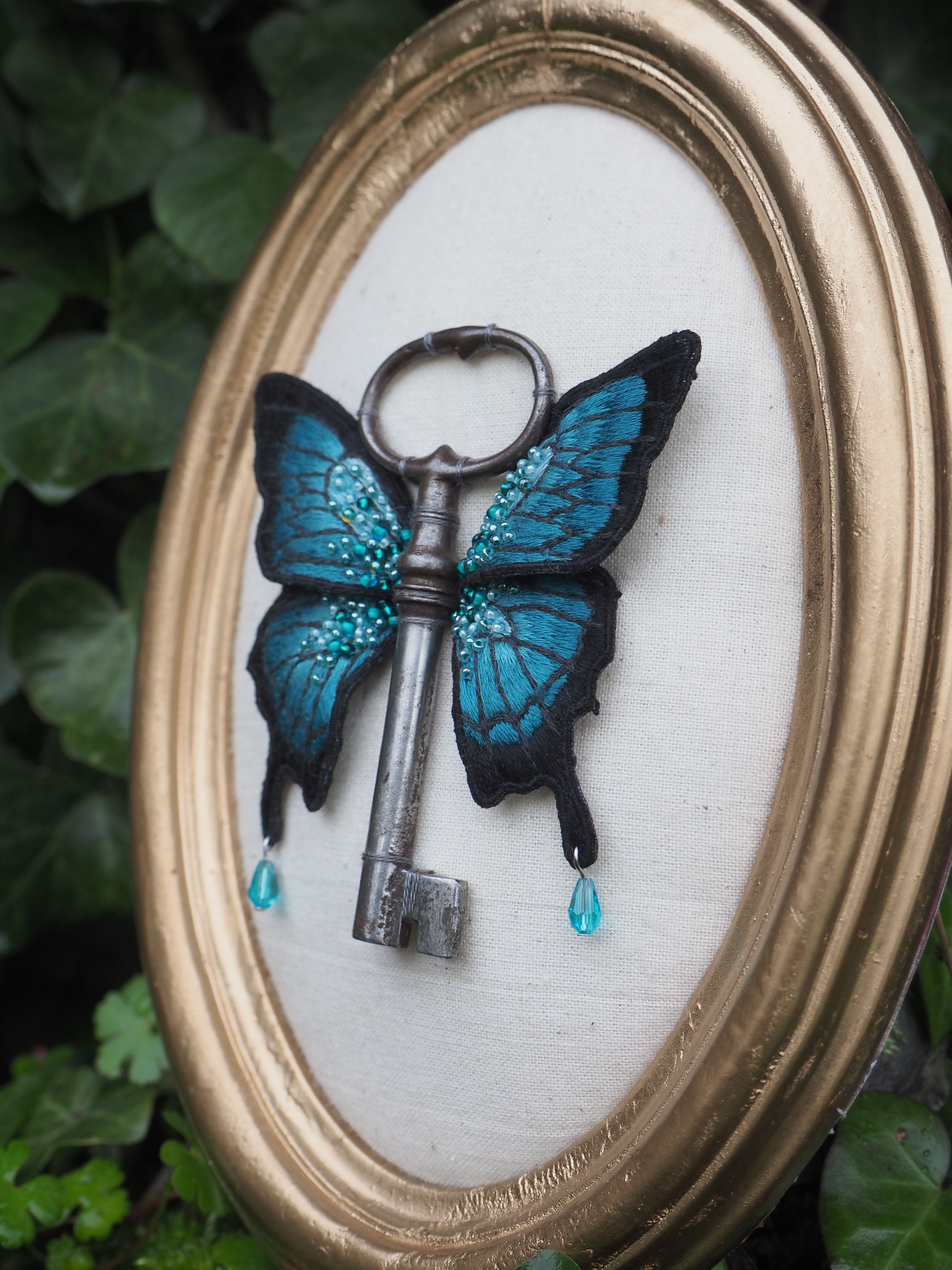 Cette image montre une création artistique très créative où une clé ancienne a été combinée avec des ailes de papillon bleues pour former un papillon-clé. L'œuvre est présentée dans un cadre ovale doré sur un fond blanc. Les ailes du papillon sont d'un bleu turquoise vibrant avec des motifs noirs, rappelant un morpho bleu. Deux petites gouttes bleues semblent tomber des ailes. La clé antique en métal sombre forme le corps du papillon. L'ensemble crée une pièce délicate et poétique qui mélange le dur et le délicat, l'ancien et le naturel. L'arrière-plan montre quelques feuilles vertes floues, suggérant que la photo a été prise dans un environnement naturel.