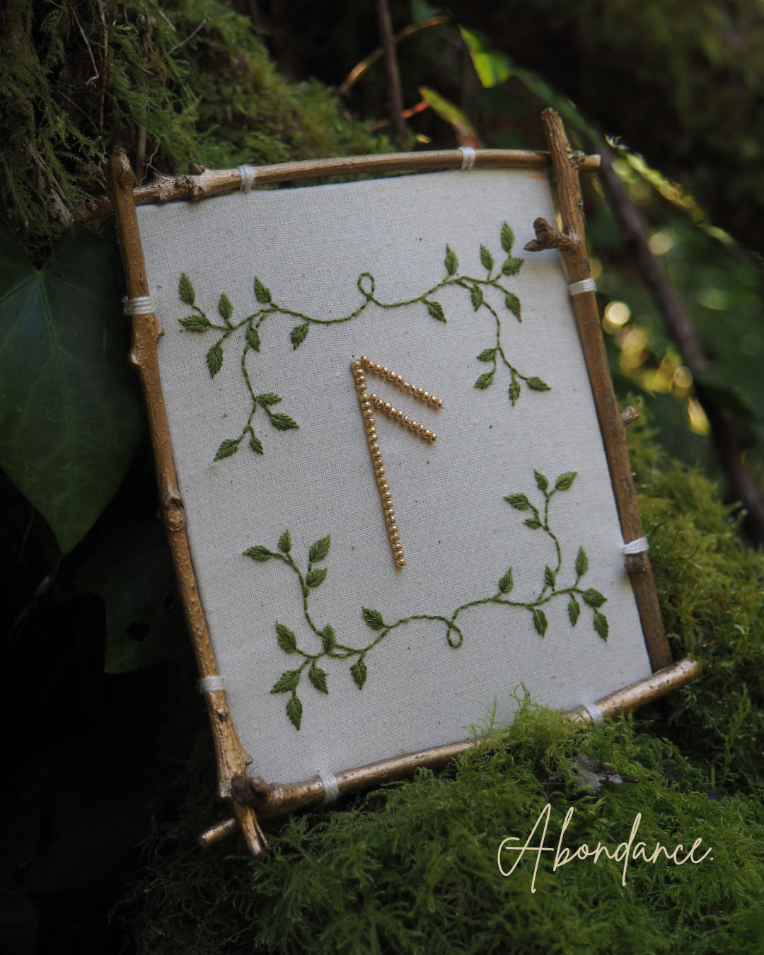 Cette image montre un symbole runique doré sur un fond blanc, encadré par un cadre rustique fait de petites branches ou de bois. Le symbole est décoré de motifs de feuilles vertes délicates qui s'entrelacent autour du cadre, créant un effet naturel et organique. Le mot "Abundance" (Abondance) est écrit en bas de l'image en écriture cursive élégante. L'ensemble est photographié sur un fond de mousse verte, donnant une ambiance très naturelle et mystique. Cette composition semble être inspirée par l'art païen ou la spiritualité nordique, où les runes sont souvent utilisées comme symboles de pouvoir ou de manifestation.