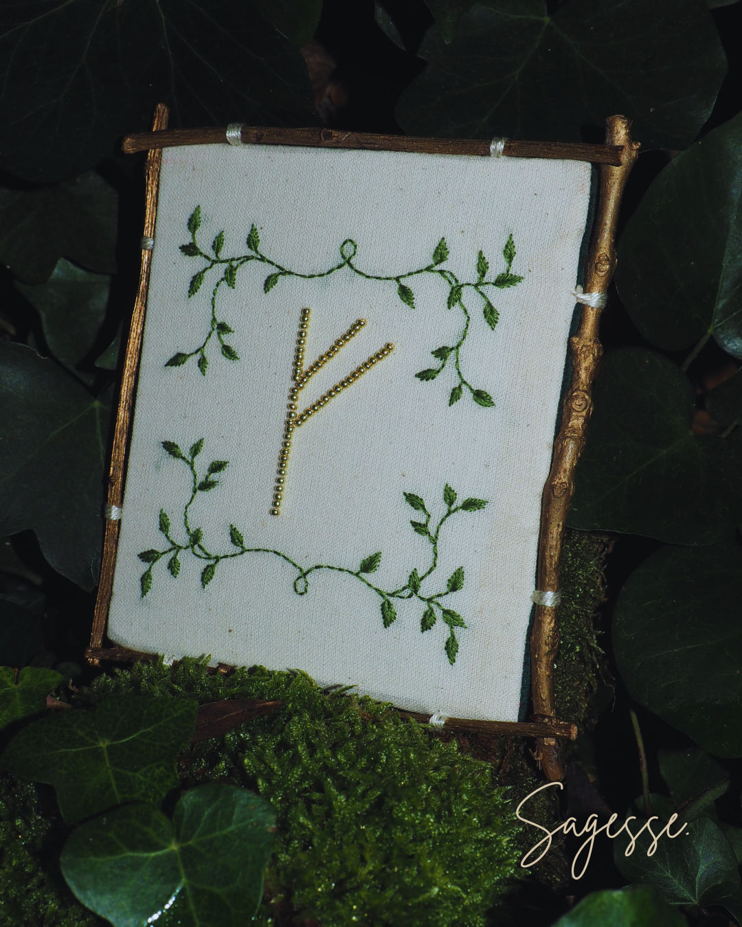 L'image présente une composition artistique et naturelle, centrée sur un cadre rustique fait de brindilles, qui encadre une pièce de tissu blanc brodée. Au premier plan, on distingue une surface recouverte de mousse verte et de feuilles de lierre, créant une base organique et forestière pour l'objet principal. Le cadre est constitué de quatre brindilles de bois, de couleur marron clair à dorée, qui sont liées aux coins pour former un rectangle. Ces brindilles ont une texture naturelle et irrégulière. À l'intérieur de ce cadre, une pièce de tissu blanc ou crème est tendue. Sur ce tissu, une broderie délicate est visible. Au centre, une rune, qui ressemble à la lettre "F" ou à la rune "Fehu", est brodée en fil doré ou jaune, avec un motif de petits points ou de perles, lui donnant un aspect scintillant. Autour de cette rune centrale, des motifs de feuilles vertes stylisées, brodées en fil vert, forment une bordure décorative, évoquant des vignes ou des branches feuillues. L'arrière-plan est flou et composé de feuillage vert foncé, probablement des feuilles de lierre ou d'autres plantes grimpantes, renforçant l'ambiance naturelle et mystique de la scène. Dans le coin inférieur droit de l'image, une signature manuscrite stylisée est visible, indiquant "Sagesse.", ajoutant une touche personnelle et artistique à la composition. L'ensemble de l'image dégage une atmosphère de calme, de nature et de spiritualité, avec une esthétique artisanale et un lien fort avec des symboles anciens ou ésotériques.