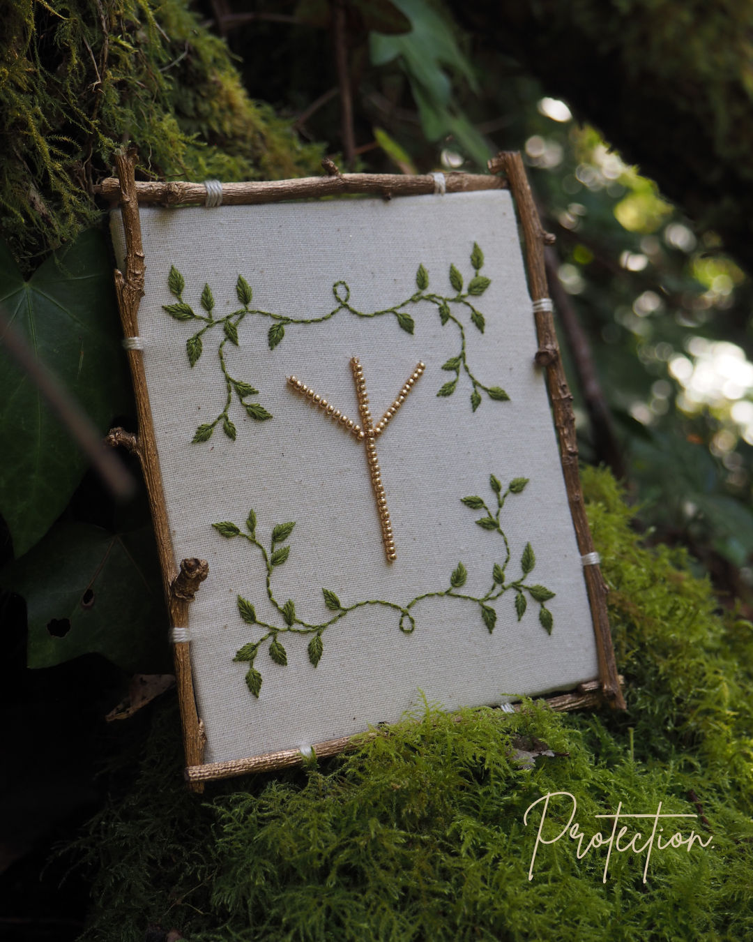 Cette image montre un symbole runique ou une rune Algiz (ᛉ) créée avec ce qui semble être des petites branches ou des brindilles, disposée sur un fond de tissu blanc ou écru. Le tissu est encadré par des branches naturelles formant un cadre rustique. Le fond est décoré d'un motif de feuilles vertes peintes ou brodées qui s'enroulent délicatement autour du symbole central. L'ensemble est photographié sur un lit de mousse verte, donnant une ambiance très naturelle et forestière. Le mot "Protection" apparaît en bas de l'image, ce qui est approprié car la rune Algiz est traditionnellement associée à la protection dans la tradition nordique. L'ensemble dégage une atmosphère païenne et naturelle très harmonieuse.