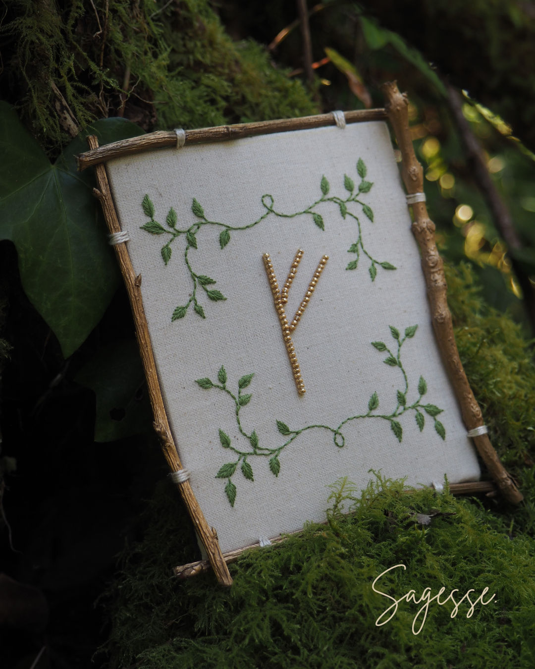 Cette image montre une belle composition artistique représentant la rune "Algiz" ou "Yr" (qui ressemble à un Y) brodée ou cousue sur un tissu blanc. Le symbole est entouré d'un motif délicat de feuilles et de vignes vertes qui s'entrelacent dans les coins. Le tissu est monté sur ce qui semble être un cadre rustique fait de petites branches ou de bois naturel. L'ensemble est photographié sur un fond de mousse verte et de feuillage, donnant une ambiance très naturelle et forestière. Le mot "Sagesse" apparaît en signature dans le coin inférieur droit de l'image. L'ensemble dégage une atmosphère très païenne, naturelle et mystique.