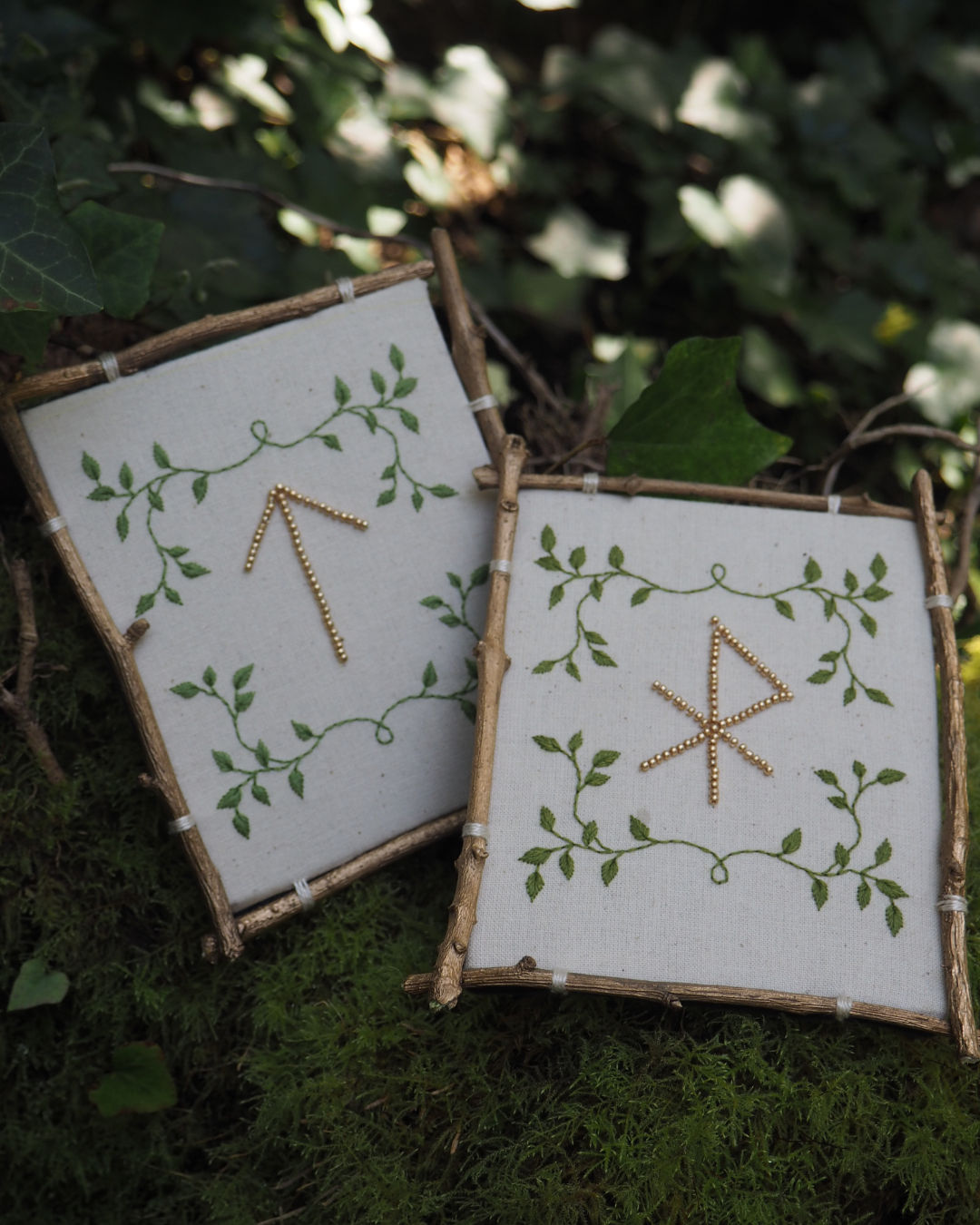 Cette image montre deux petits panneaux ou cadres artisanaux faits avec des branches de bois naturel. Sur chaque panneau blanc, on peut voir des motifs décoratifs : des feuilles vertes délicates qui forment une bordure végétale, et au centre de chaque panneau, des symboles runiques ou mystiques créés avec ce qui semble être du fil doré ou métallique. Les panneaux sont photographiés sur un fond de mousse verte, donnant une ambiance naturelle et mystique à l'ensemble. Le style est très rustique et évoque un artisanat inspiré de la nature ou des traditions païennes.