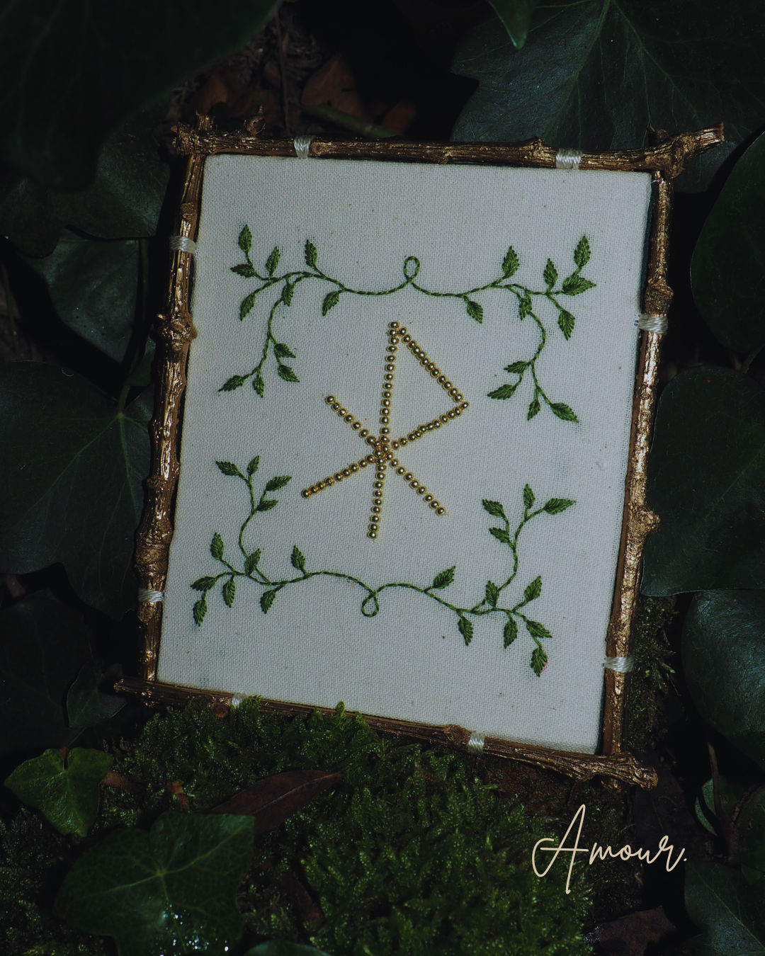 L'image présente une broderie encadrée, posée sur un lit de mousse et de feuilles de lierre, créant une ambiance naturelle et mystique. Au centre de l'image, un cadre de forme carrée ou légèrement rectangulaire, dont les bords semblent être faits de petites branches ou de brindilles dorées, contient une broderie sur un tissu de couleur crème ou blanc cassé. La broderie représente un motif central qui ressemble à une rune ou un symbole ancien, formé de lignes droites et obliques, réalisées avec des perles dorées ou des fils brillants. Autour de ce symbole central, des motifs de feuilles et de vrilles vertes sont brodés, encadrant le symbole et ajoutant une touche organique. Le cadre est posé sur un sol recouvert de mousse verte luxuriante et de feuilles de lierre vert foncé, qui s'étendent sur les côtés et en arrière-plan. La lumière semble douce et diffuse, créant des ombres subtiles et mettant en valeur les textures des éléments naturels. Dans le coin inférieur droit de l'image, le mot "Amour" est écrit en cursive dorée ou jaune pâle, ajoutant une dimension émotionnelle ou thématique à la scène. L'ensemble de l'image dégage une atmosphère de calme, de nature et de spiritualité, avec une touche d'élégance rustique.