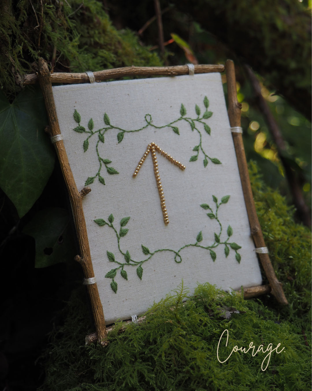 Cette image montre une création artisanale délicate : un symbole de rune ou une flèche pointant vers le haut, réalisée avec ce qui semble être des petites perles ou des bâtonnets dorés. Ce motif est encadré par des branches vertes brodées ou peintes sur un fond blanc. Le tout est présenté dans un cadre rustique fait de petites branches naturelles assemblées. L'œuvre est photographiée sur un lit de mousse verte, donnant une ambiance très naturelle et forestière à l'ensemble. Le mot "Courage" apparaît en écriture cursive dans le coin inférieur droit de l'image. L'ensemble dégage une atmosphère paisible et inspirante, mêlant éléments naturels et artisanat délicat.