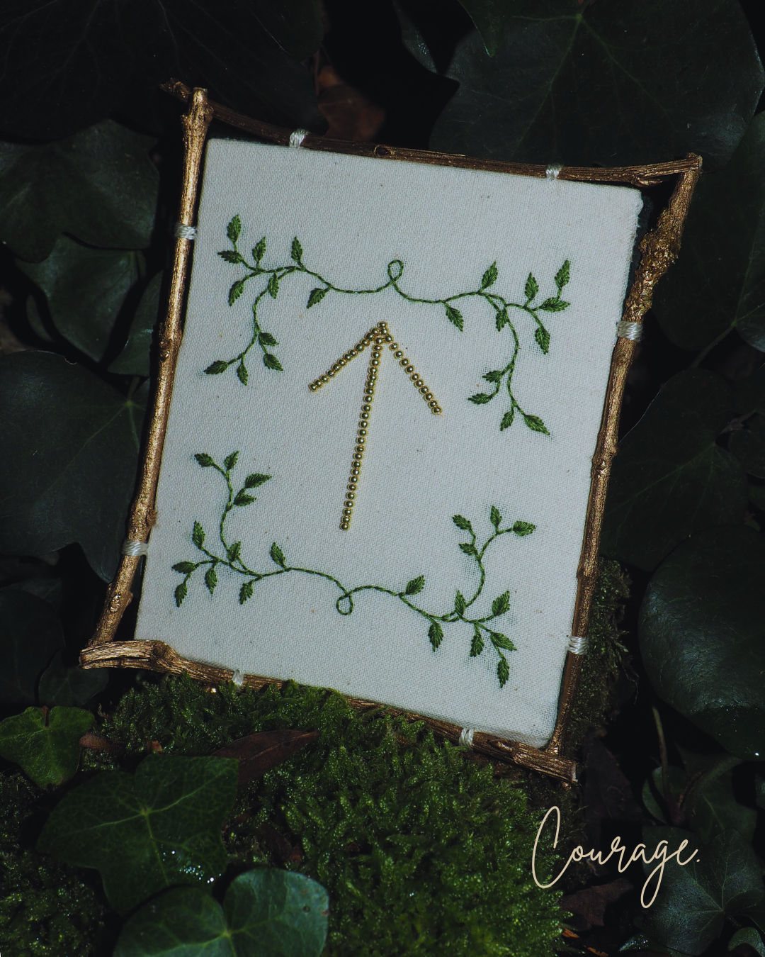 L'image présente une broderie encadrée, posée sur un lit de mousse et de feuilles de lierre, créant une ambiance naturelle et forestière. Au centre de l'image, un cadre rustique, probablement fait de brindilles ou de petits morceaux de bois dorés, encadre une pièce de tissu de couleur crème ou blanc cassé. Sur ce tissu, une broderie délicate est visible. Le motif principal est une flèche pointant vers le haut, brodée avec des perles ou des fils dorés, lui donnant un aspect scintillant. Autour de cette flèche, des motifs de feuilles vertes sont brodés, formant des guirlandes ou des arabesques qui encadrent la flèche, renforçant le thème végétal. Le cadre est posé sur un tapis de mousse verte luxuriante, parsemé de feuilles de lierre vert foncé, certaines avec des reflets brillants. L'arrière-plan est flou, mais on distingue des feuillages sombres, suggérant un environnement boisé ou un jardin. Dans le coin inférieur droit de l'image, le mot "Courage" est écrit en lettres cursives dorées ou de couleur claire, ajoutant une signification ou un message à la scène. L'éclairage semble doux et naturel, mettant en valeur les textures de la mousse, des feuilles et la brillance de la broderie dorée. L'ensemble de l'image dégage une atmosphère de calme, de nature et de symbolisme.