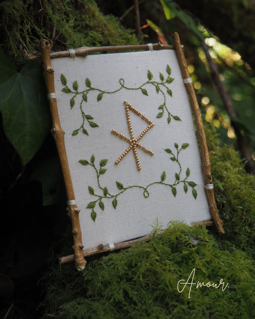 Cette image montre un ouvrage de broderie encadré dans un cadre rustique fait de petites branches ou bâtons de bois. La broderie est réalisée sur un tissu blanc et présente un motif central qui ressemble à une rune ou un symbole nordique fait de points dorés ou de fils métalliques. Ce symbole est entouré d'une délicate broderie de feuilles et de tiges vertes qui forment un motif végétal décoratif aux quatre coins. L'ensemble est photographié sur un fond naturel de mousse et de verdure, créant une ambiance féerique et forestière. Le mot "Amour" apparaît en signature dans le coin inférieur droit de l'image.