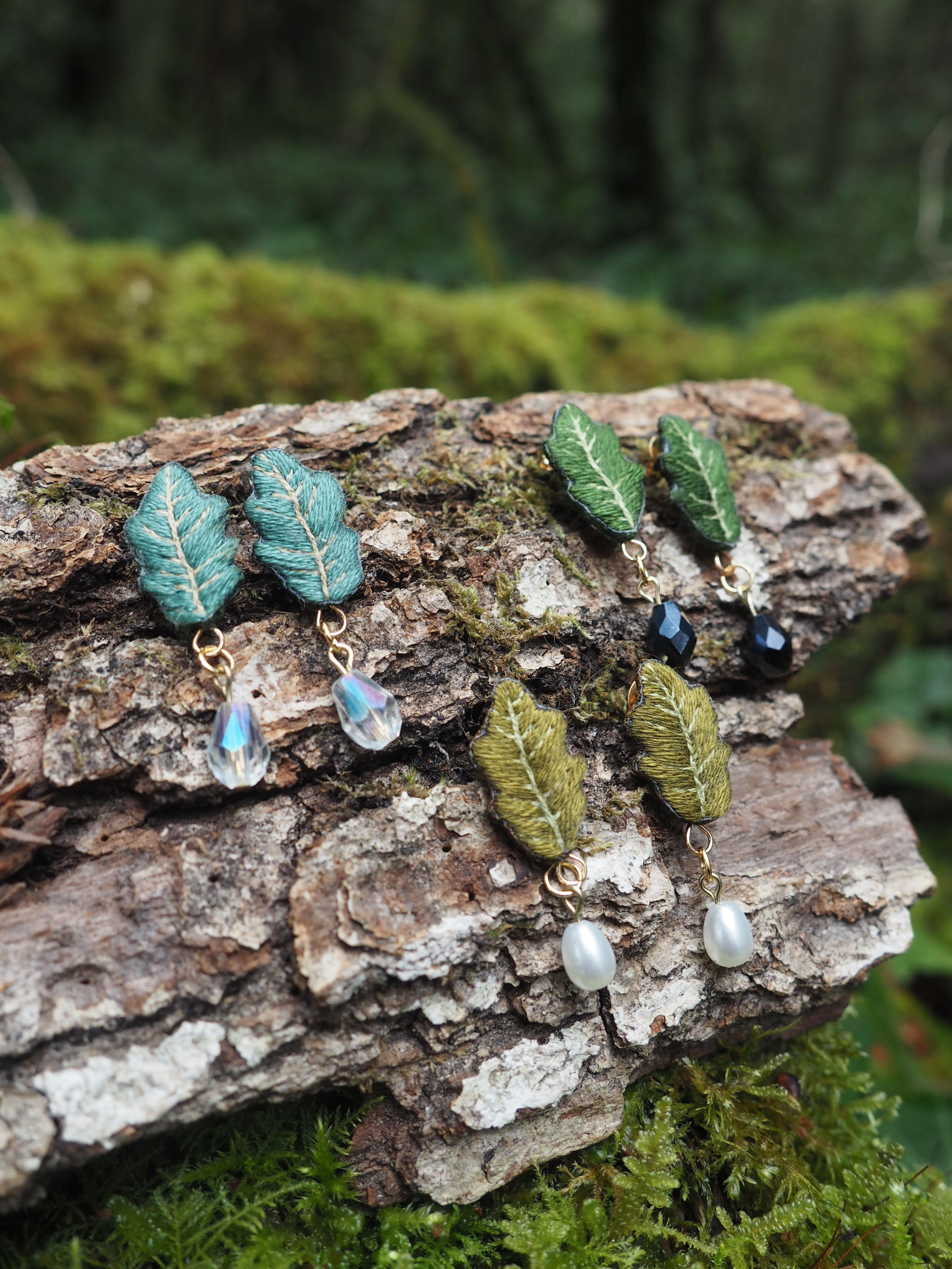 L'image présente quatre paires de boucles d'oreilles artisanales, chacune conçue pour ressembler à des feuilles, posées sur un morceau de bois brut et texturé, recouvert de mousse verte. L'arrière-plan est flou, suggérant un environnement forestier ou naturel. Voici une description détaillée des boucles d'oreilles et de leur environnement : **Les Boucles d'Oreilles :**
Chaque boucle d'oreille est composée d'une petite feuille brodée ou texturée, suspendue à un crochet d'oreille (non visible sur toutes les boucles, mais implicite par leur nature). Sous chaque feuille pend une perle ou une breloque. 1.  **Paire en haut à gauche :** Deux boucles d'oreilles avec des feuilles de couleur vert-bleu ou sarcelle. Elles ont une texture nervurée qui imite les feuilles naturelles. Sous chaque feuille pend une perle en forme de goutte, transparente et facettée, qui semble capter la lumière. Les attaches sont dorées. 2.  **Paire en haut à droite :** Deux boucles d'oreilles avec des feuilles d'un vert plus foncé, presque émeraude. La texture est similaire aux précédentes. Sous chaque feuille pend une perle ronde et foncée, probablement noire ou bleu nuit, qui contraste avec la couleur de la feuille. Les attaches sont dorées. 3.  **Paire en bas à gauche :** Deux boucles d'oreilles avec des feuilles d'un vert olive ou kaki. La texture est également nervurée. Sous chaque feuille pend une perle de forme irrégulière, de couleur crème ou blanche, qui ressemble à une perle de culture naturelle. Les attaches sont dorées. 4.  **Paire en bas à droite :** Il semble s'agir de la même paire que celle en bas à gauche, mais elle est légèrement décalée, confirmant la présence de deux boucles d'oreilles identiques. **Le Support :**
Les boucles d'oreilles sont disposées sur un morceau de bois mort, probablement une branche ou un tronc tombé. Le bois est de couleur marron foncé, avec une écorce rugueuse et des fissures, ce qui lui confère une texture rustique. Des taches de mousse verte, de différentes nuances, recouvrent partiellement le bois, ajoutant à l'ambiance naturelle. **L'Arrière-plan :**
L'arrière-plan est un flou artistique (bokeh) de verdure, composé de feuilles et de mousse, dans des tons de vert variés. Cela renforce l'idée que les boucles d'oreilles sont présentées dans un cadre naturel, évoquant la forêt ou un jardin luxuriant. La lumière semble douce et diffuse, typique d'un sous-bois. **Ambiance Générale :**
L'image dégage une atmosphère calme et naturelle, mettant en valeur le caractère artisanal et organique des bijoux. Les couleurs sont harmonieuses, dominées par les verts et les bruns, avec des touches de couleurs plus vives apportées par les perles.