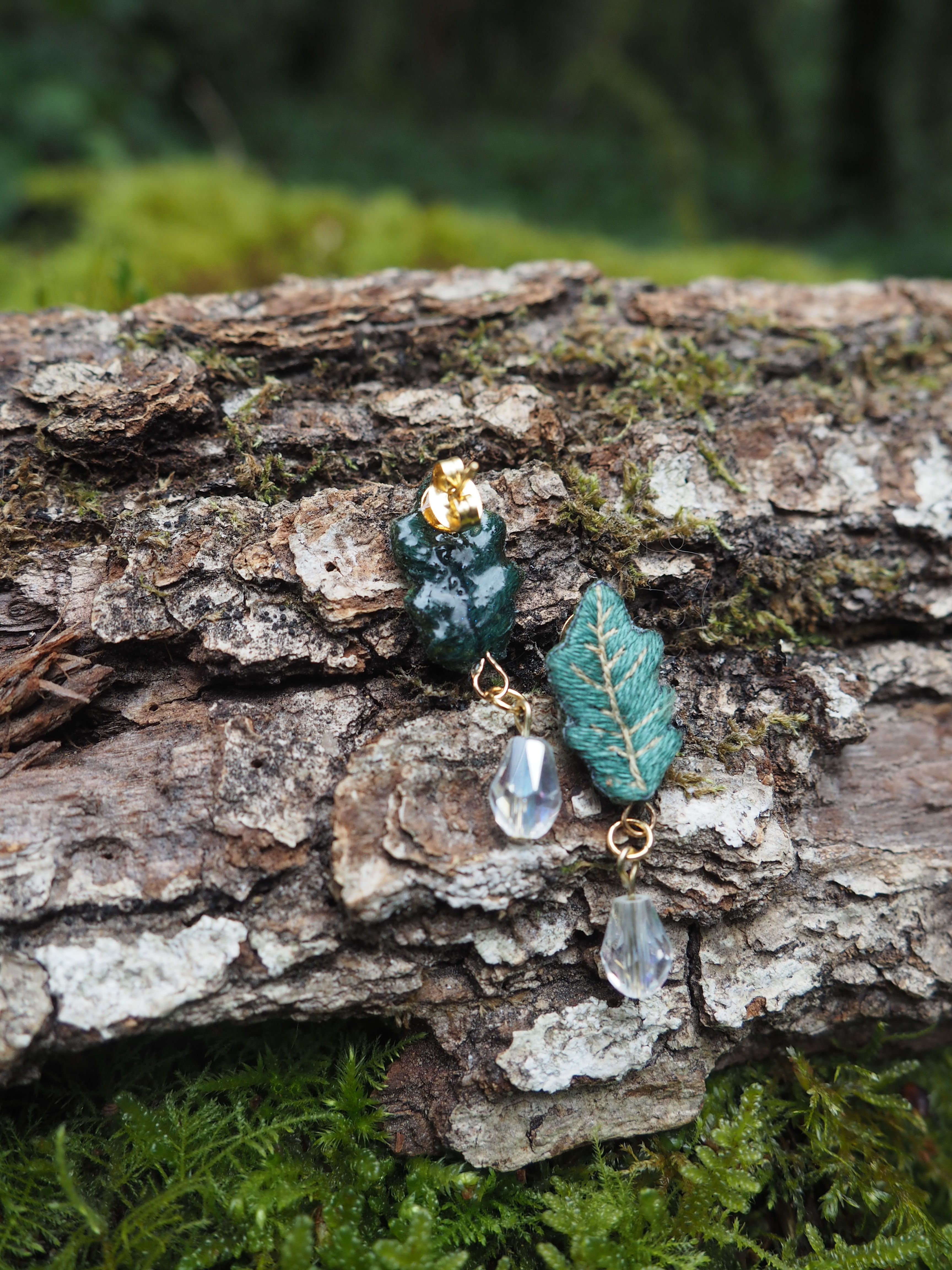 L'image présente une paire de boucles d'oreilles fantaisie, posées sur un morceau de bois brut et texturé, recouvert de mousse. **Description des boucles d'oreilles :**
*   **Matériaux et Couleurs :** Les boucles d'oreilles sont composées de plusieurs éléments. La partie supérieure de chaque boucle d'oreille est de couleur vert foncé, avec une texture qui pourrait évoquer une feuille ou une forme organique. Elles sont surmontées d'un petit élément doré, probablement l'attache ou une perle décorative.
*   **Pendentifs :** Sous ces éléments verts, des pendentifs sont suspendus par de petits anneaux dorés. Chaque pendentif est une perle transparente et facettée, qui semble capter et réfléchir la lumière, donnant un éclat cristallin.
*   **Design :** Le design général est délicat et semble inspiré par la nature, avec des couleurs et des formes qui rappellent la forêt. L'une des boucles d'oreilles, celle de droite, présente un élément vert plus clairement identifiable comme une feuille, avec des nervures apparentes. **Description de l'arrière-plan :**
*   **Support :** Les boucles d'oreilles reposent sur un morceau de bois mort, dont l'écorce est rugueuse et crevassée, avec des zones plus claires et plus foncées. De la mousse verte et du lichen sont visibles sur le bois, ajoutant à l'ambiance naturelle.
*   **Environnement :** L'arrière-plan est flou, mais on distingue des teintes de vert, suggérant un environnement forestier ou végétal, probablement de la mousse ou des fougères. La profondeur de champ est faible, ce qui met l'accent sur les boucles d'oreilles. **Lumière et Ambiance :**
La lumière semble naturelle et douce, éclairant les boucles d'oreilles et faisant scintiller les perles transparentes. L'ambiance générale est calme, naturelle et met en valeur le caractère artisanal ou "fait main" des bijoux.