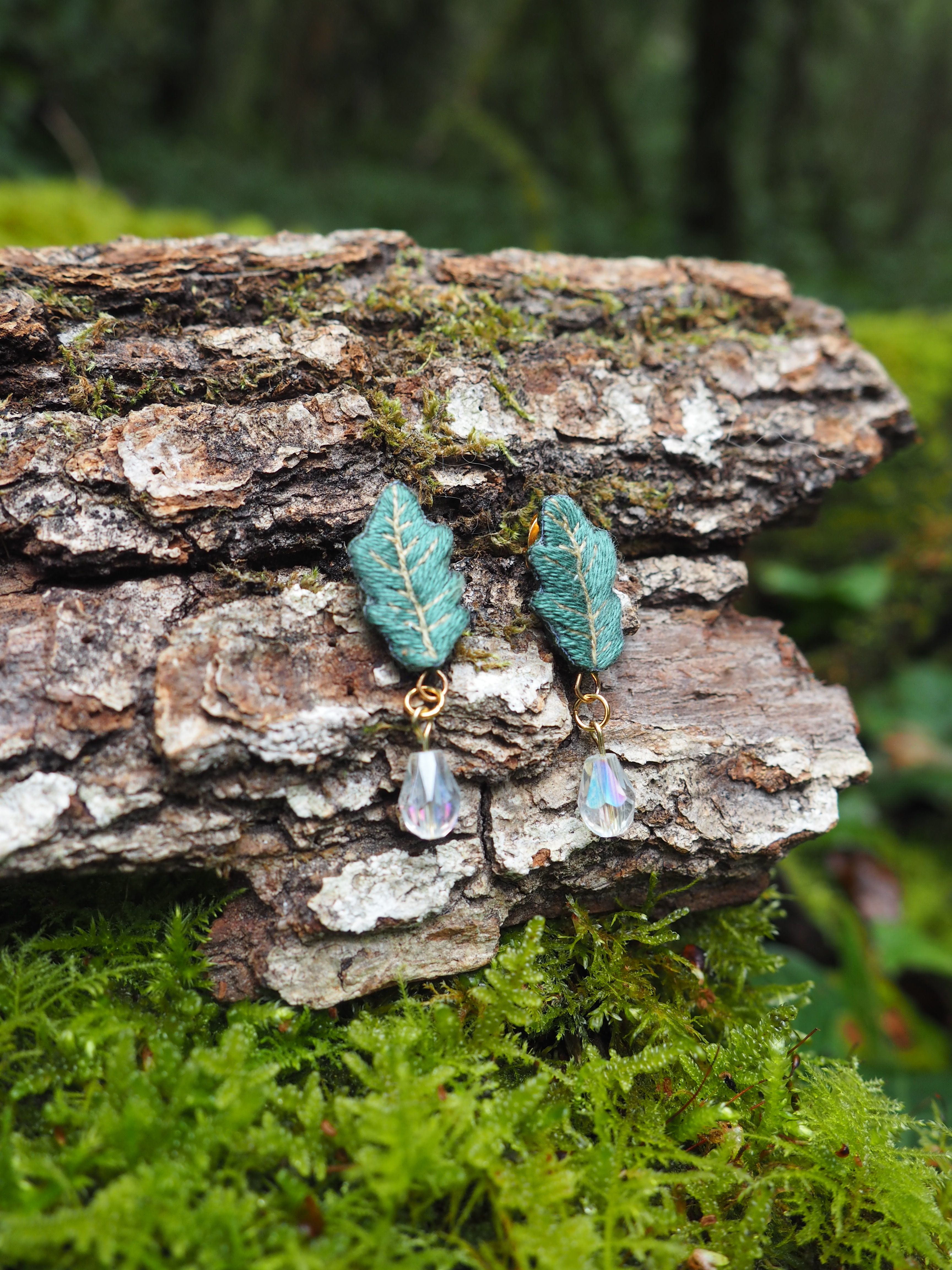 L'image présente une paire de boucles d'oreilles pendantes, posées sur un morceau d'écorce d'arbre texturée, recouverte de mousse verte. Chaque boucle d'oreille est composée d'un motif en forme de feuille de chêne stylisée, de couleur vert-bleu ou sarcelle, avec des détails de nervures apparents, suggérant une texture brodée ou sculptée. Sous chaque feuille pend une petite perle transparente et facettée, qui semble capter la lumière, ajoutant une touche d'éclat. Les attaches des boucles d'oreilles sont de couleur dorée. L'arrière-plan est flou, mais on distingue des tons de vert et de brun, indiquant un environnement naturel, probablement une forêt ou un sous-bois, ce qui complète le thème naturel des boucles d'oreilles. La lumière semble douce et diffuse, mettant en valeur les détails des bijoux et la texture de l'écorce et de la mousse.
