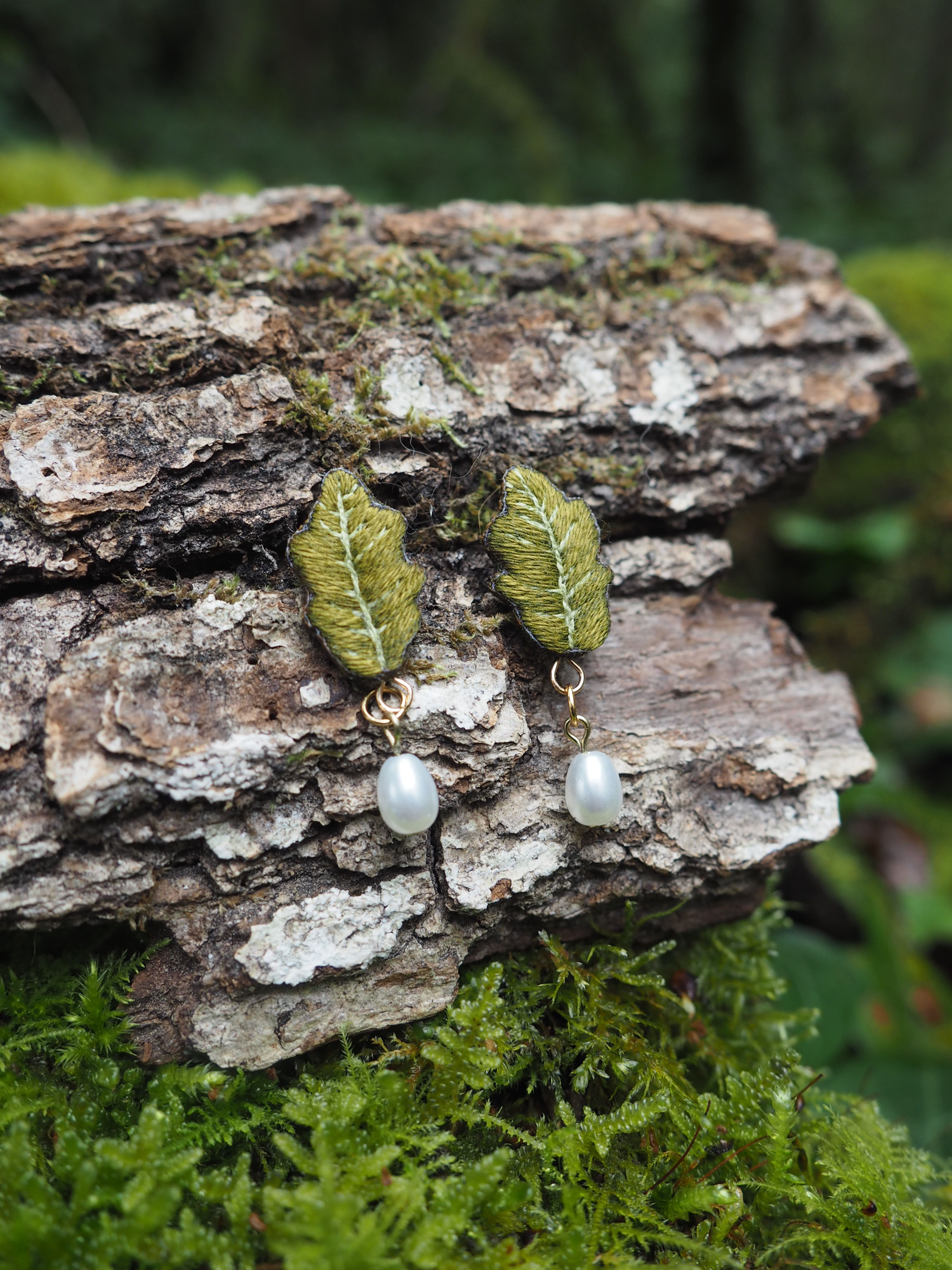 L'image présente une paire de boucles d'oreilles délicates et artisanales, posées sur un morceau d'écorce d'arbre texturée et recouverte de mousse. **Description des boucles d'oreilles :**
Chaque boucle d'oreille est composée de deux éléments principaux :
1.  **La partie supérieure (feuille) :** Il s'agit d'une forme de feuille stylisée, brodée avec du fil vert olive ou kaki. La broderie est fine, avec des lignes qui imitent les nervures d'une feuille, donnant un aspect naturel et organique. La texture de la broderie est visible, ajoutant de la profondeur à l'objet.
2.  **La partie inférieure (perle) :** Sous chaque feuille brodée pend une perle de culture de couleur blanc crème ou ivoire. Les perles sont de forme irrégulière, ce qui suggère qu'elles sont naturelles ou d'imitation réaliste, et ont un éclat doux et nacré. Les boucles d'oreilles sont reliées par de petits anneaux métalliques dorés, qui ajoutent une touche d'élégance et de chaleur. **Description de l'arrière-plan et du support :**
Les boucles d'oreilles sont posées sur un morceau d'écorce d'arbre, probablement un tronc ou une branche coupée. L'écorce est très texturée, avec des crevasses et des reliefs prononcés, et des nuances de brun et de gris. Une fine couche de mousse verte, d'un vert plus vif que les feuilles des boucles d'oreilles, recouvre certaines parties de l'écorce et le fond de l'image, renforçant l'ambiance naturelle et forestière. **Lumière et ambiance :**
La lumière semble naturelle et diffuse, éclairant bien les boucles d'oreilles et mettant en valeur leurs détails. L'ambiance générale de l'image est douce, naturelle et évoque la nature, l'artisanat et la délicatesse. En résumé, l'image met en scène des boucles d'oreilles uniques, inspirées par la nature, avec une esthétique rustique et élégante, présentées dans un cadre qui souligne leur origine et leur design organique.