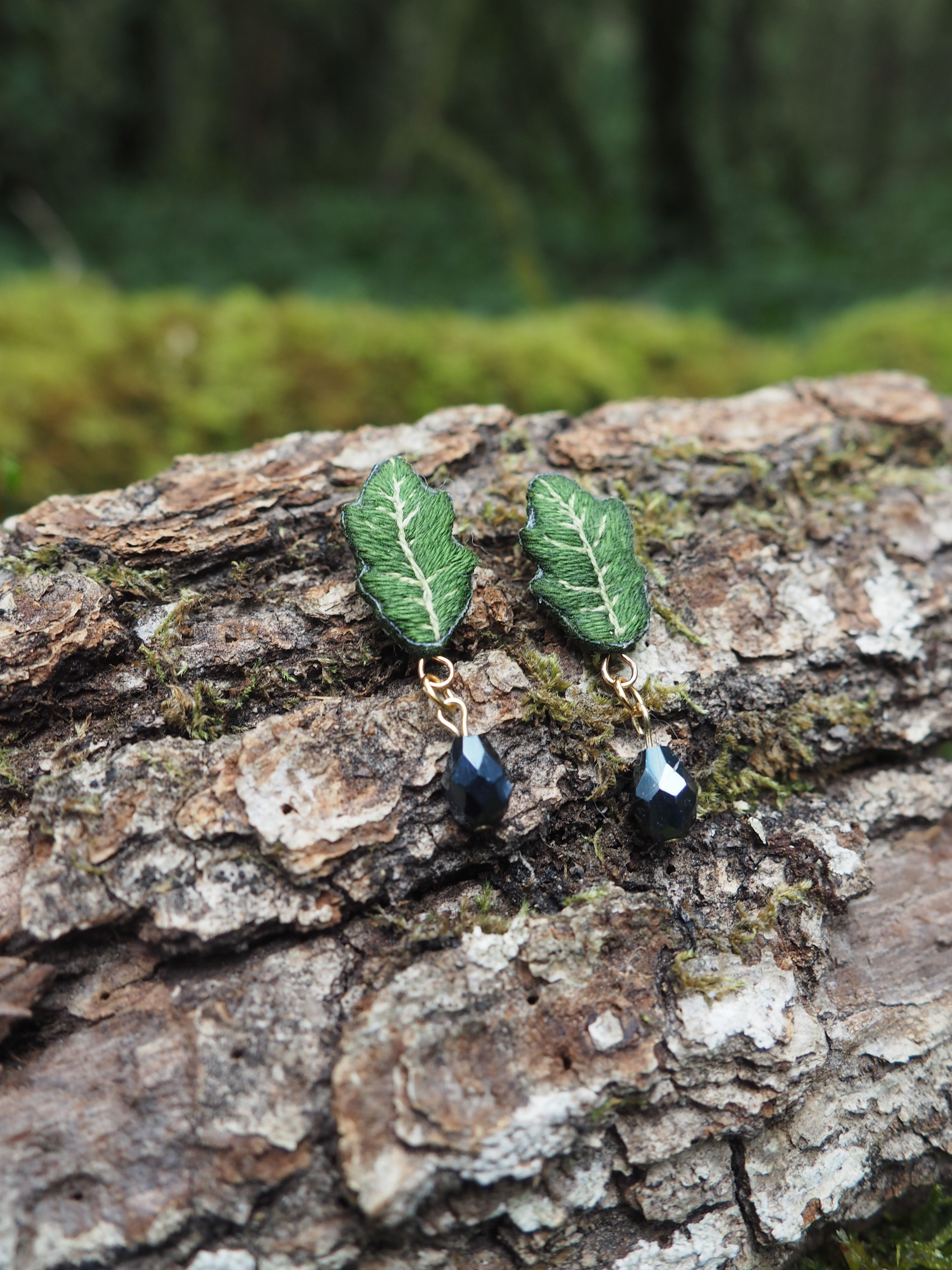 L'image présente une paire de boucles d'oreilles artisanales posées sur un morceau d'écorce d'arbre. **Description des boucles d'oreilles :**
*   Chaque boucle d'oreille est composée d'un motif supérieur en forme de feuille verte. Ces feuilles ont une texture détaillée, avec des nervures blanches ou très claires qui contrastent avec le vert foncé de la feuille, leur donnant un aspect réaliste et texturé.
*   Sous chaque feuille, pend une petite perle ou un cristal de couleur sombre, probablement noir ou gris foncé, qui capte la lumière et ajoute une touche d'éclat.
*   Les éléments sont reliés par de petits anneaux ou tiges métalliques de couleur dorée, qui ajoutent une touche d'élégance. **Contexte et arrière-plan :**
*   Les boucles d'oreilles sont posées sur un morceau d'écorce d'arbre rugueuse et texturée, de couleur marron clair à grisâtre, avec des crevasses et des aspérités. On peut apercevoir de petites touches de mousse verte sur l'écorce, renforçant le thème naturel.
*   L'arrière-plan est flou, mais on distingue des teintes de vert et de marron, suggérant un environnement forestier ou végétal, ce qui complète parfaitement le design des boucles d'oreilles en forme de feuille. **Ambiance générale :**
L'image dégage une ambiance naturelle, artisanale et délicate. La mise au point est nette sur les boucles d'oreilles, les mettant en valeur comme des objets uniques et faits à la main, inspirés par la nature.