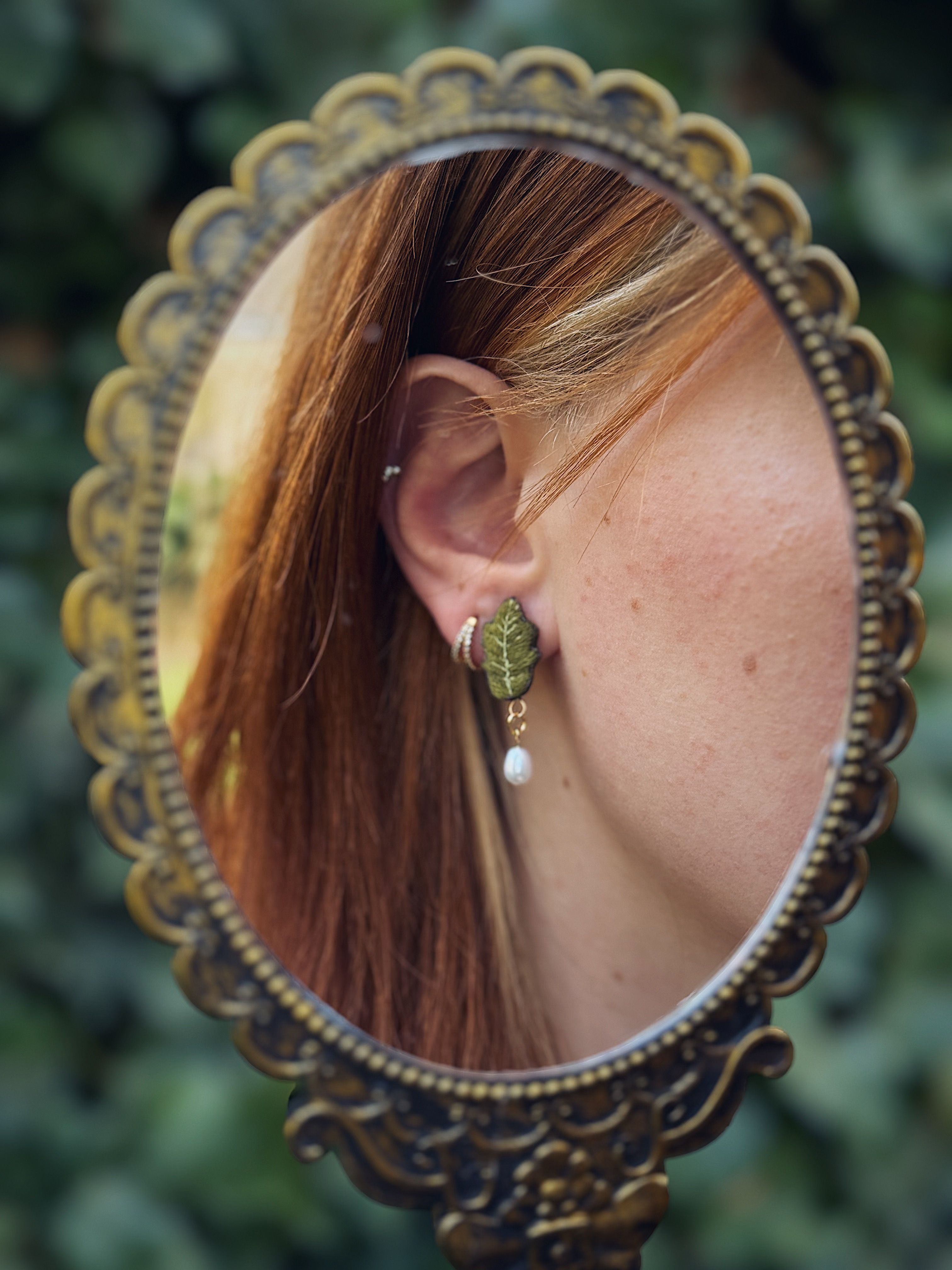 Cette image présente un gros plan d'une oreille de femme, reflétée dans un miroir de style vintage. Voici une description détaillée des propriétés visuelles : **Le miroir :**
*   Le miroir est de forme ovale et possède un cadre orné de couleur bronze ou laiton vieilli. Les détails du cadre suggèrent un style ancien ou baroque, avec des motifs en relief.
*   Le miroir est tenu ou posé de manière à refléter l'oreille de la personne. **L'oreille et le bijou :**
*   L'oreille est celle d'une femme, avec une peau claire parsemée de quelques taches de rousseur.
*   Les cheveux de la personne sont roux ou châtain clair avec des reflets roux, et ils encadrent l'oreille.
*   Un piercing discret est visible sur le cartilage supérieur de l'oreille.
*   La personne porte une boucle d'oreille pendante. Le motif principal de la boucle d'oreille est une petite feuille verte, détaillée et texturée, qui semble être faite d'un matériau comme l'argile polymère ou le métal émaillé. Sous la feuille, pend une petite perle blanche ou crème, de forme légèrement irrégulière, ce qui lui donne un aspect naturel ou baroque. **L'arrière-plan (hors du miroir) :**
*   L'arrière-plan visible autour du miroir est flou, mais on peut distinguer des teintes vertes, suggérant la présence de feuillage ou de végétation, comme des plantes ou des buissons. Cela crée un contraste doux avec le cadre du miroir et met en valeur le sujet principal. **L'éclairage et l'ambiance :**
*   L'éclairage semble naturel et doux, peut-être en extérieur ou près d'une fenêtre, ce qui met en valeur les détails de la boucle d'oreille et la texture de la peau.
*   L'ambiance générale de l'image est délicate, naturelle et un peu nostalgique, accentuée par le style du miroir et le motif de la boucle d'oreille.