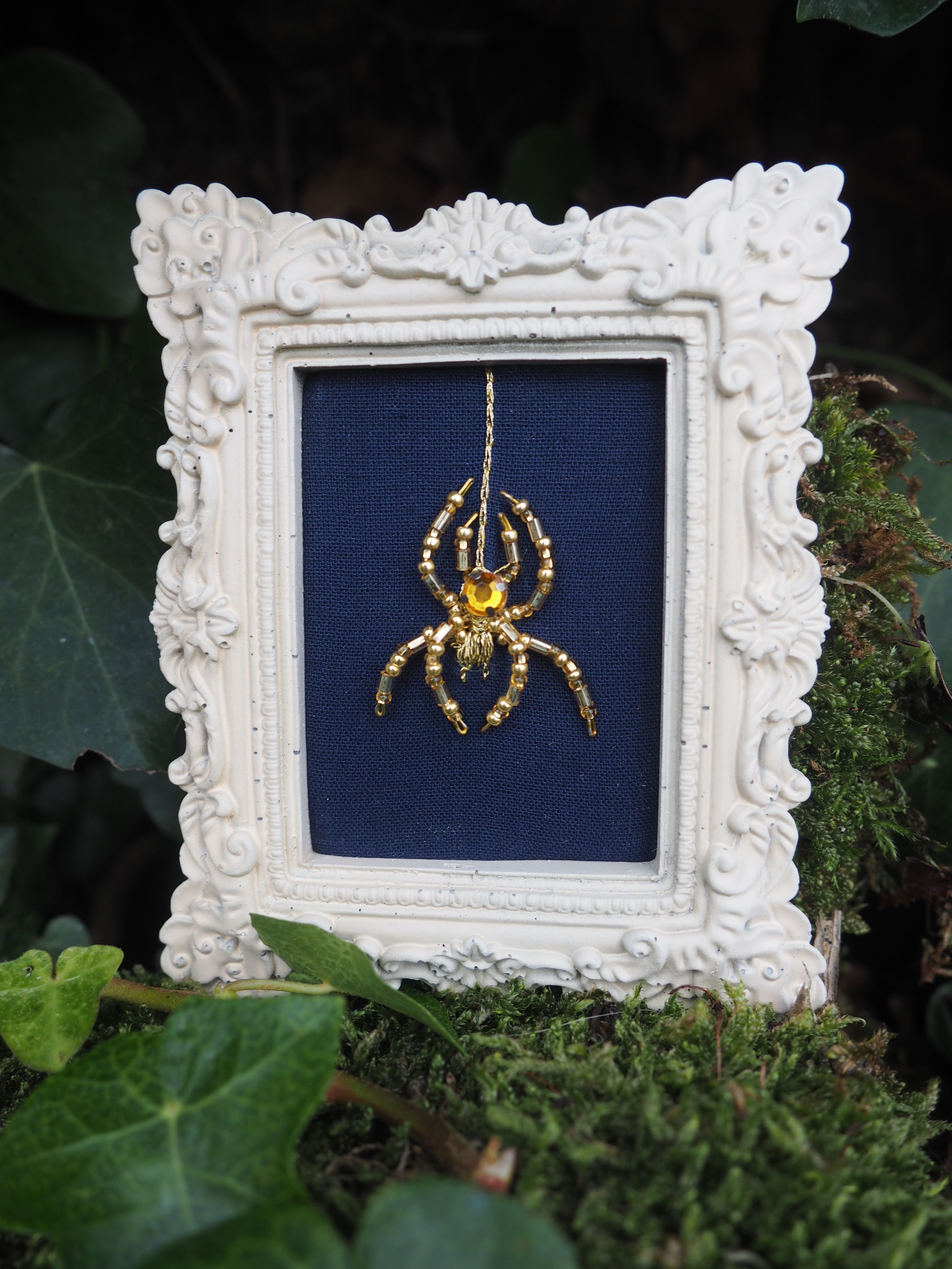 Cette image présente une œuvre d'art décorative encadrée, photographiée dans un environnement naturel verdoyant.  **Le cadre** : Il s'agit d'un cadre baroque blanc orné de motifs sculptés élaborés, avec des volutes, des fleurs et des détails décoratifs en relief qui lui donnent un aspect très ornemental et classique. **L'œuvre** : À l'intérieur du cadre se trouve une broderie ou un travail textile sur fond bleu marine. Le motif central représente une araignée dorée, probablement brodée au fil d'or ou doré, qui se détache élégamment sur le tissu sombre. **L'environnement** : L'ensemble est photographié posé sur de la mousse verte, entouré de feuillage et de végétation, créant un contraste saisissant entre l'objet d'art raffiné et l'environnement naturel sauvage. Cette mise en scène crée une atmosphère mystérieuse et poétique, mêlant l'artisanat délicat à la nature, avec l'araignée dorée comme symbole central qui fait écho à l'environnement naturel tout en étant sublimée par l'art textile.