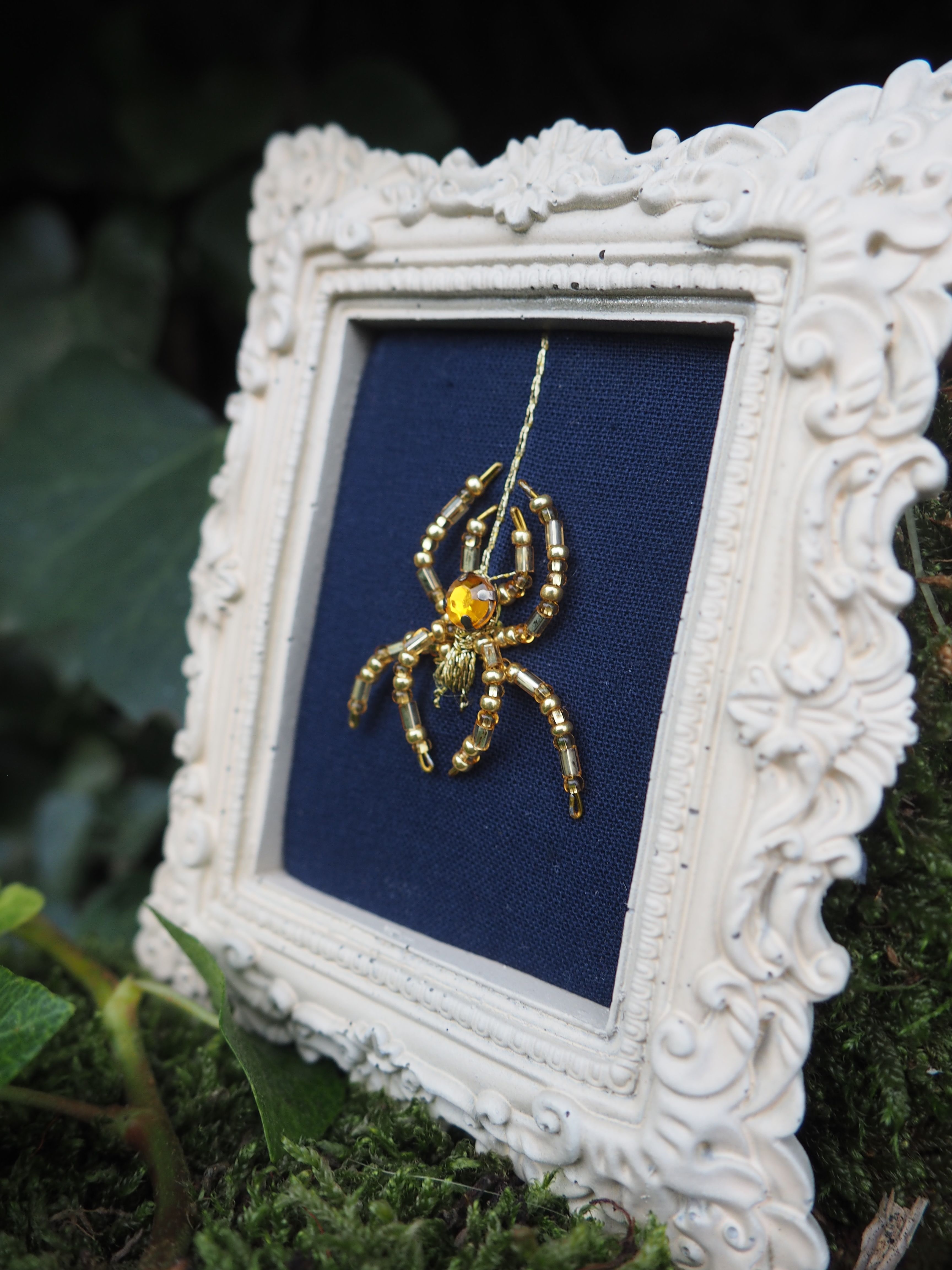 Cette image présente une œuvre d'art encadrée de manière très originale. Il s'agit d'une araignée dorée brodée ou tissée sur un fond de tissu bleu marine. L'araignée est représentée avec un grand détail, montrant ses huit pattes et son corps segmenté dans des tons dorés brillants qui contrastent magnifiquement avec le fond sombre. Le cadre est particulièrement remarquable : il s'agit d'un cadre baroque blanc orné de motifs décoratifs en relief, avec des volutes et des ornementations sculptées qui lui donnent un aspect très élégant et sophistiqué. L'ensemble est photographié dans un environnement naturel, posé sur ce qui semble être de la mousse ou de la végétation verte, créant un contraste intéressant entre l'œuvre d'art raffinée et son environnement naturel. Cette mise en scène souligne peut-être le lien entre l'art et la nature, l'araignée étant elle-même un élément du monde naturel représenté de manière artistique. L'éclairage met bien en valeur les détails dorés de la broderie et la texture du cadre blanc, créant une composition visuellement très attrayante.