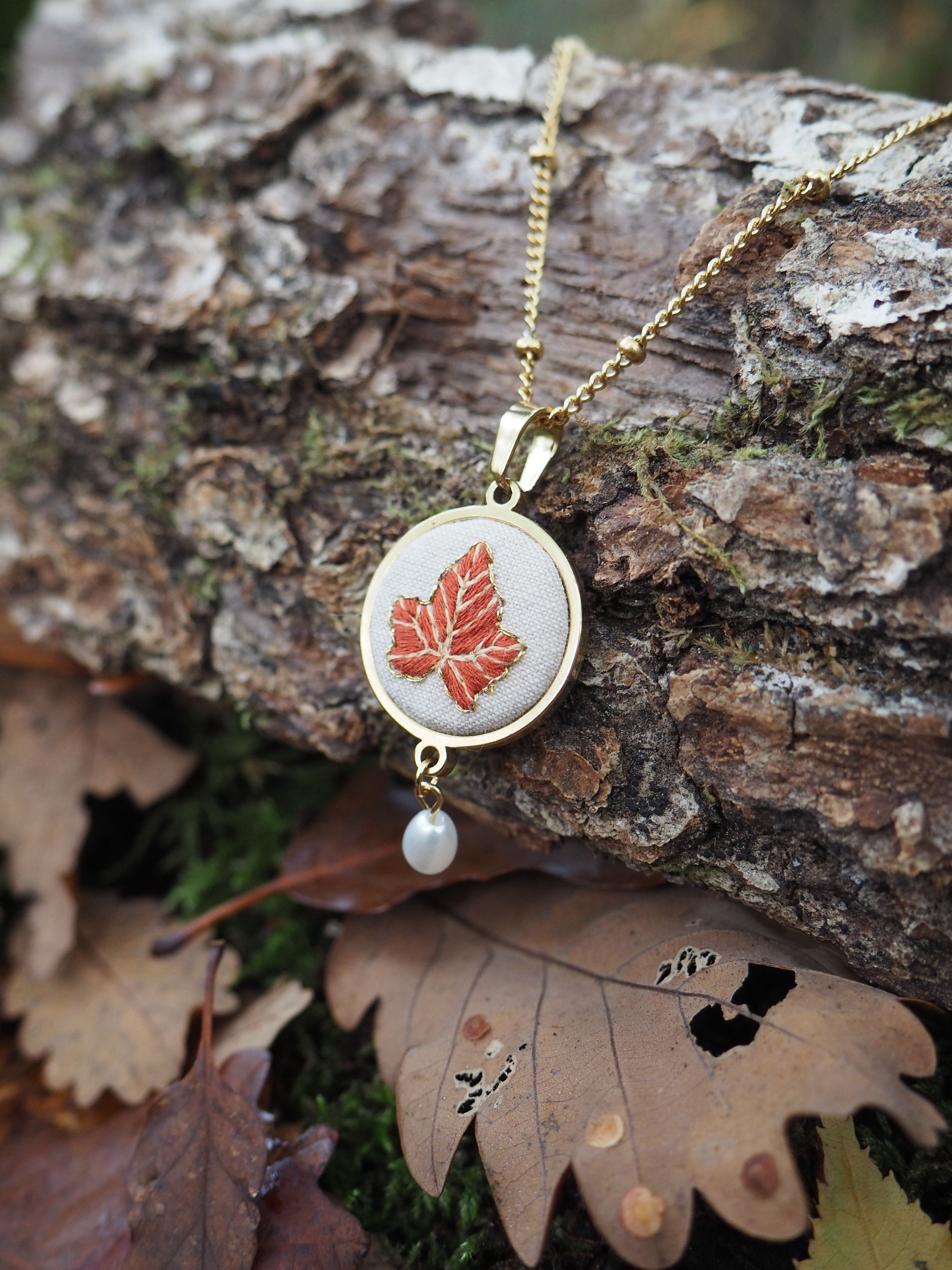 Cette image présente un élégant collier disposé sur un tronc d'arbre dans un cadre naturel automnal. Le bijou se compose d'un pendentif circulaire à monture dorée contenant un motif de feuille d'érable rouge brodée ou imprimée sur un fond blanc ou crème. Une petite perle blanche nacrée pend délicatement sous le médaillon, ajoutant une touche de raffinement. Le collier repose sur une chaîne dorée fine qui serpente sur l'écorce rugueuse et texturée du tronc. L'environnement naturel est parfaitement en harmonie avec le thème du bijou : des feuilles d'automne aux teintes brunes et dorées jonchent le sol autour du tronc, créant une mise en scène poétique qui évoque la saison automnale. L'éclairage naturel met en valeur les détails du pendentif et crée un contraste saisissant entre les tons chauds du bijou et la palette terrestre de l'environnement forestier. Cette composition suggère un style de bijouterie artisanale ou vintage, célébrant la beauté de la nature à travers un accessoire délicat et symbolique.