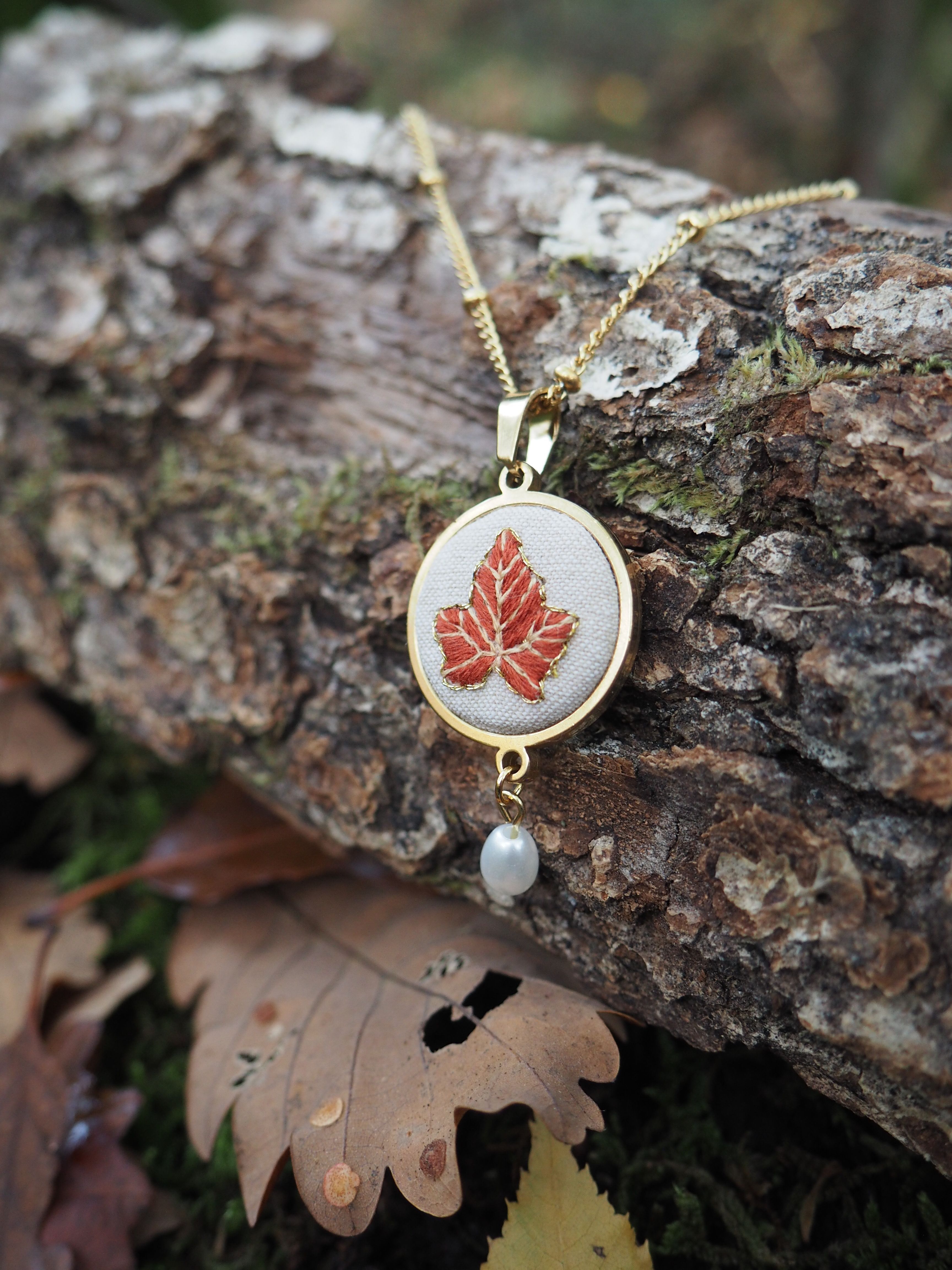 Cette image présente un élégant collier posé sur une bûche de bois dans un environnement naturel automnal. Le bijou se compose d'un pendentif circulaire avec un cadre doré qui encadre un motif de feuille d'érable rouge brodée ou imprimée sur un fond blanc ou crème. Une petite perle blanche pend délicatement sous le pendentif principal. Le collier repose sur une chaîne dorée fine qui serpente sur l'écorce rugueuse et texturée du bois. L'arrière-plan flou révèle des feuilles d'automne tombées au sol, créant une harmonie parfaite entre le motif de la feuille d'érable du bijou et l'environnement naturel qui l'entoure. La photographie capture magnifiquement les tons chauds de l'automne - les dorés du métal, les rouges de la feuille d'érable, et les bruns naturels du bois et des feuilles mortes. Cette mise en scène évoque la beauté saisonnière et donne au bijou une dimension poétique et naturelle.