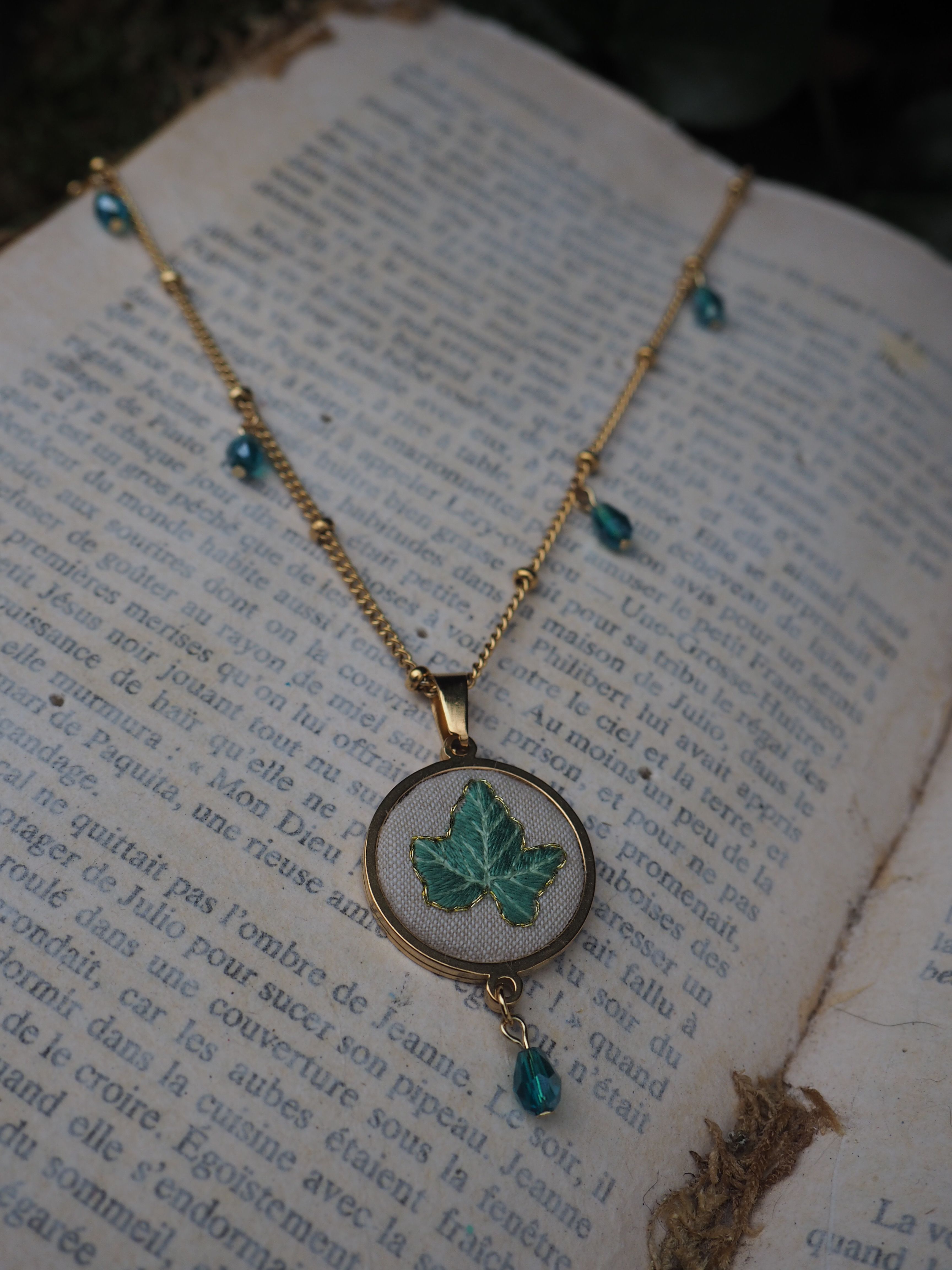 This image features a close-up of a delicate gold-toned necklace resting on an open book with aged, yellowed pages. The necklace has a thin chain adorned with small, evenly spaced gold beads and several small, faceted teal or dark blue beads that dangle at intervals along its length. The central pendant is oval-shaped, with a gold-toned frame. Inside the frame, against a light, possibly white or cream-colored fabric background, is an embroidered or painted design of a single ivy leaf in shades of green. A single matching teal/dark blue faceted bead dangles from the bottom of the oval pendant. The book's pages are filled with printed text in French, suggesting an old novel or literary work. The lighting is soft, highlighting the necklace and the texture of the paper, with a slightly darker, out-of-focus background that hints at natural elements like foliage. The overall aesthetic is vintage, natural, and somewhat whimsical.