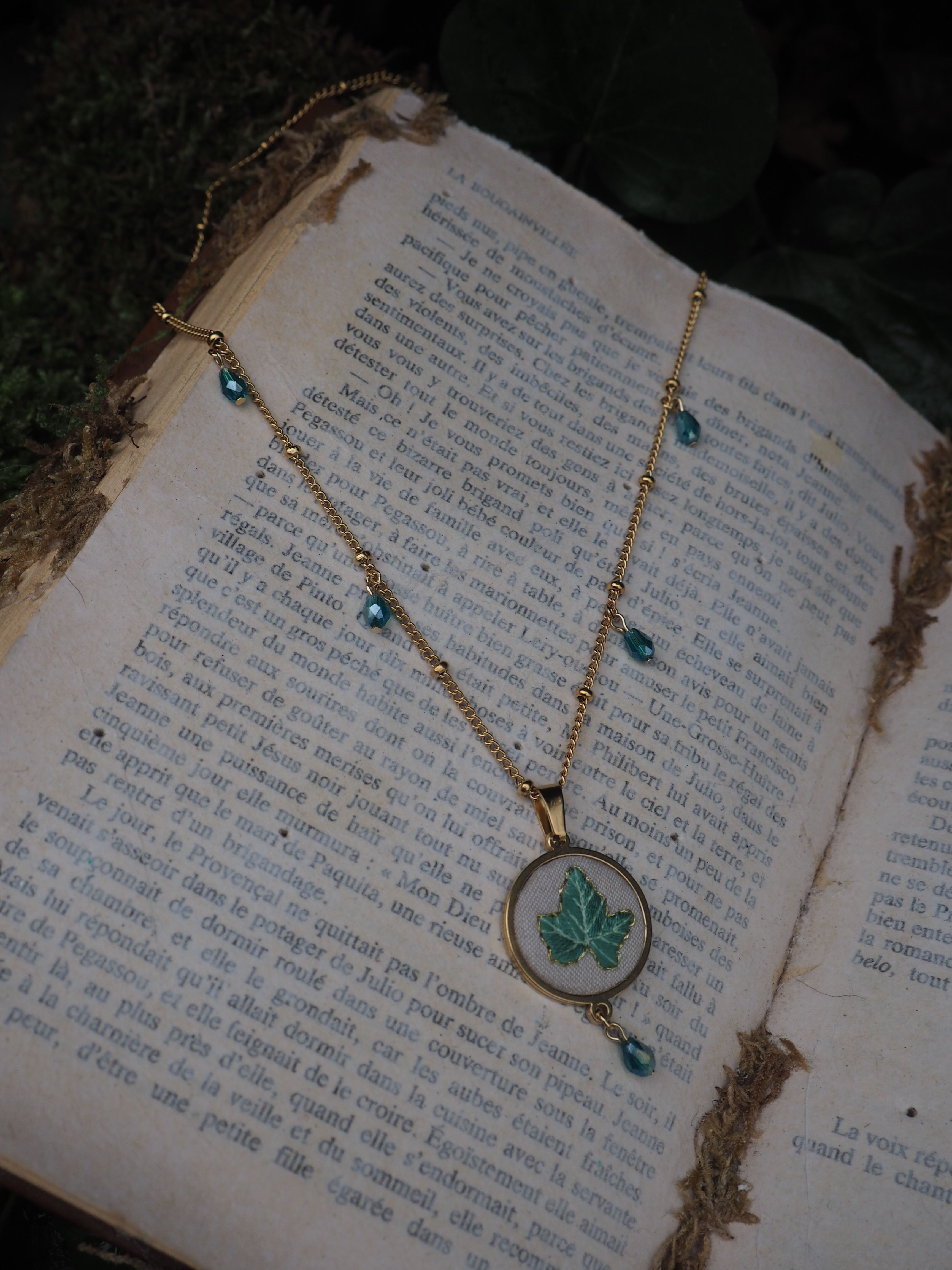 This image features a gold necklace with a pendant and blue beads, resting on an open book. The necklace is made of a delicate gold chain with small gold beads interspersed along its length. Several small, faceted blue beads are attached to the chain at intervals, adding a touch of color. The central pendant is circular, with a gold frame, and features a light green leaf design on a white background. Another blue bead dangles from the bottom of the pendant. The necklace is laid out on an open book with aged, yellowed pages. The book is filled with text printed in black ink, suggesting an old or vintage publication. The pages appear somewhat worn, with slight discoloration and perhaps some minor fraying at the edges. The background of the image is softly blurred, showing hints of dark green foliage or moss, which contributes to a natural, slightly rustic, or antique aesthetic. The lighting appears soft, highlighting the necklace and the book.