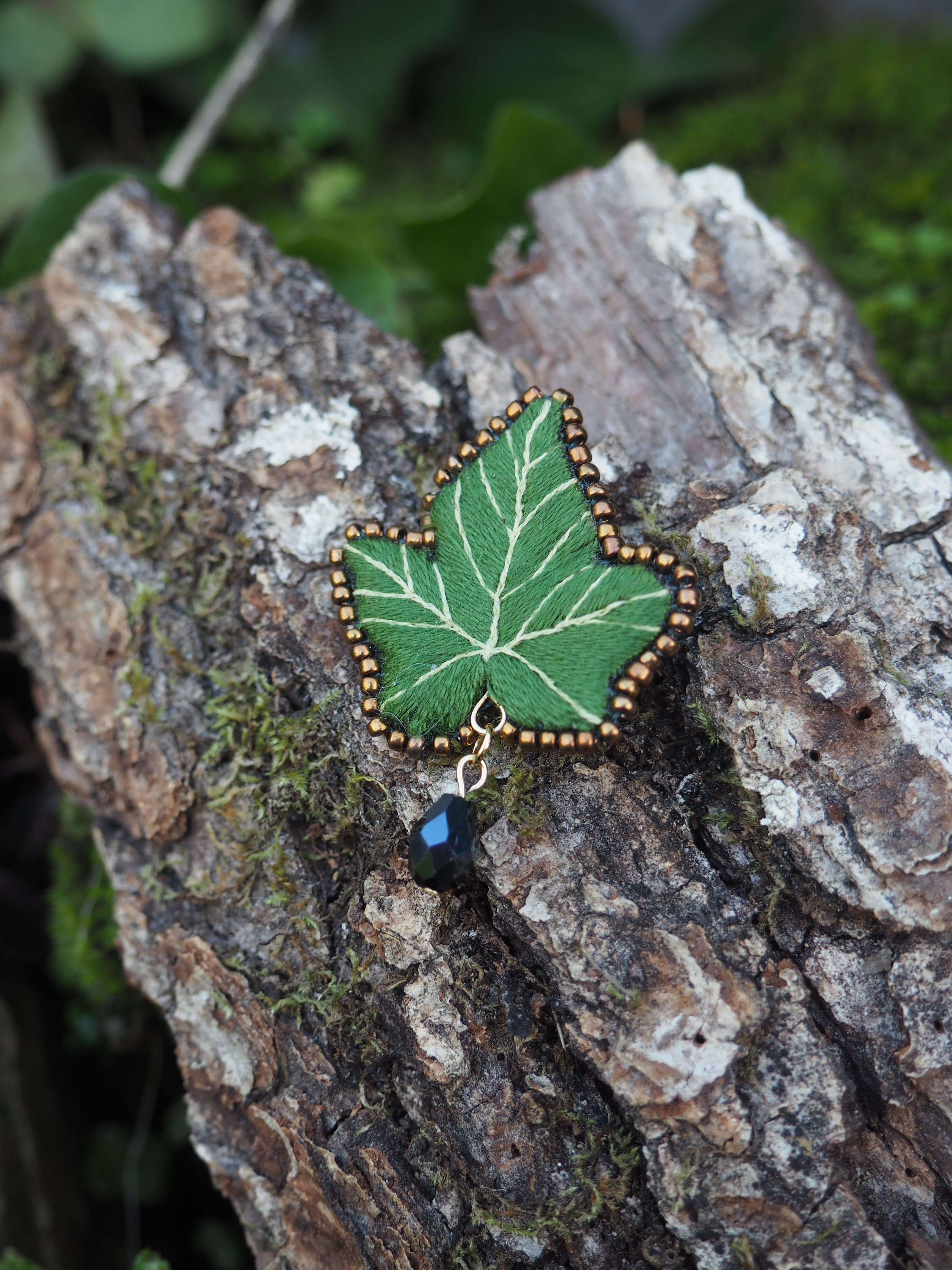 L'image présente une broche en forme de feuille de lierre, posée sur un morceau d'écorce d'arbre. **Description de la broche :**
*   La broche est de couleur verte, imitant la teinte d'une feuille de lierre.
*   Elle est brodée avec des fils plus clairs pour représenter les nervures de la feuille, ajoutant du réalisme et du détail.
*   Le contour de la feuille est orné de petites perles dorées ou de points de broderie de couleur or, ce qui lui donne une finition élégante et un léger éclat.
*   Sous la broche, suspendue par un petit anneau doré, se trouve une perle noire ou un petit cristal taillé, qui ajoute un élément de pendeloque et de sophistication. **Contexte et arrière-plan :**
*   La broche est posée sur un morceau d'écorce d'arbre, qui est rugueuse et texturée, avec des nuances de brun et de gris.
*   De la mousse verte est visible par endroits sur l'écorce, suggérant un environnement naturel et humide.
*   L'arrière-plan est flou, mais on peut distinguer des feuillages verts, ce qui renforce l'idée d'une scène en extérieur ou d'un cadre naturel.
*   La lumière semble douce et naturelle, mettant en valeur les détails de la broche sans créer d'ombres dures. L'ensemble de l'image dégage une atmosphère artisanale et naturelle, soulignant la beauté et la délicatesse de la broche faite à la main.
